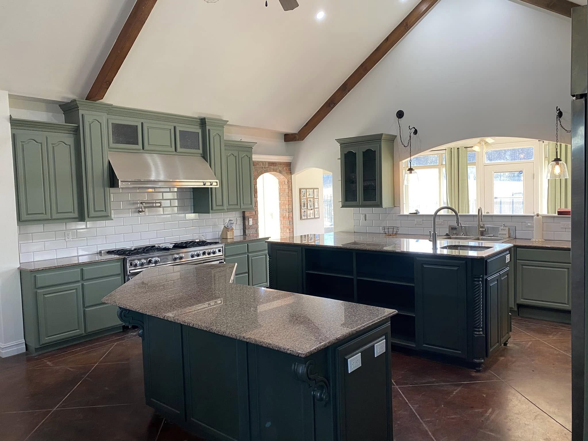 A spacious kitchen with green cabinets, a black kitchen island, and white subway tile backsplash. There are stainless steel appliances, a large window with green curtains, and a high, vaulted ceiling with wooden beams.