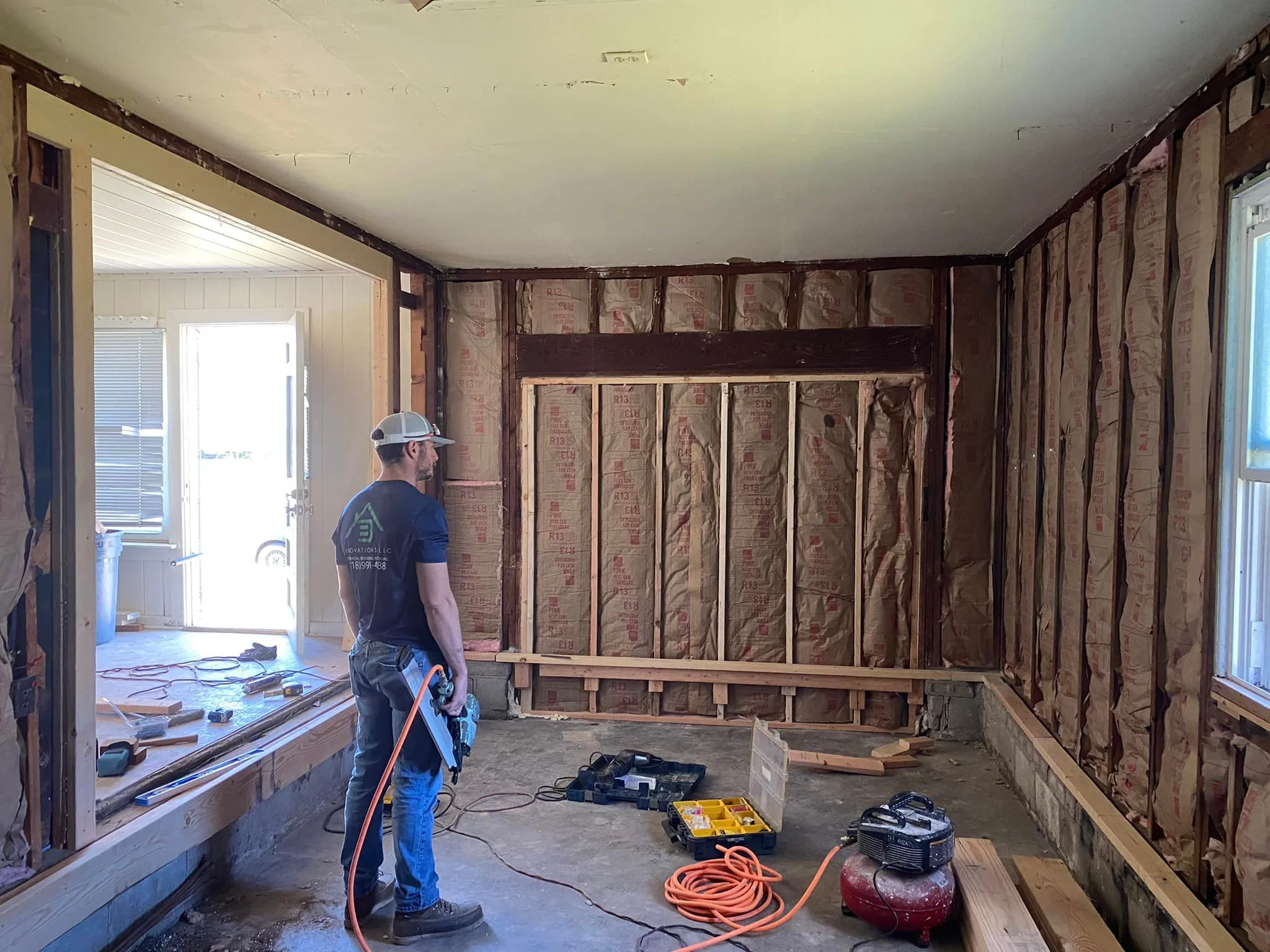 A man in a baseball cap standing in a room under construction, holding an air compressor hose, with insulation exposed on the walls, construction tools on the floor, and a doorway leading outside.