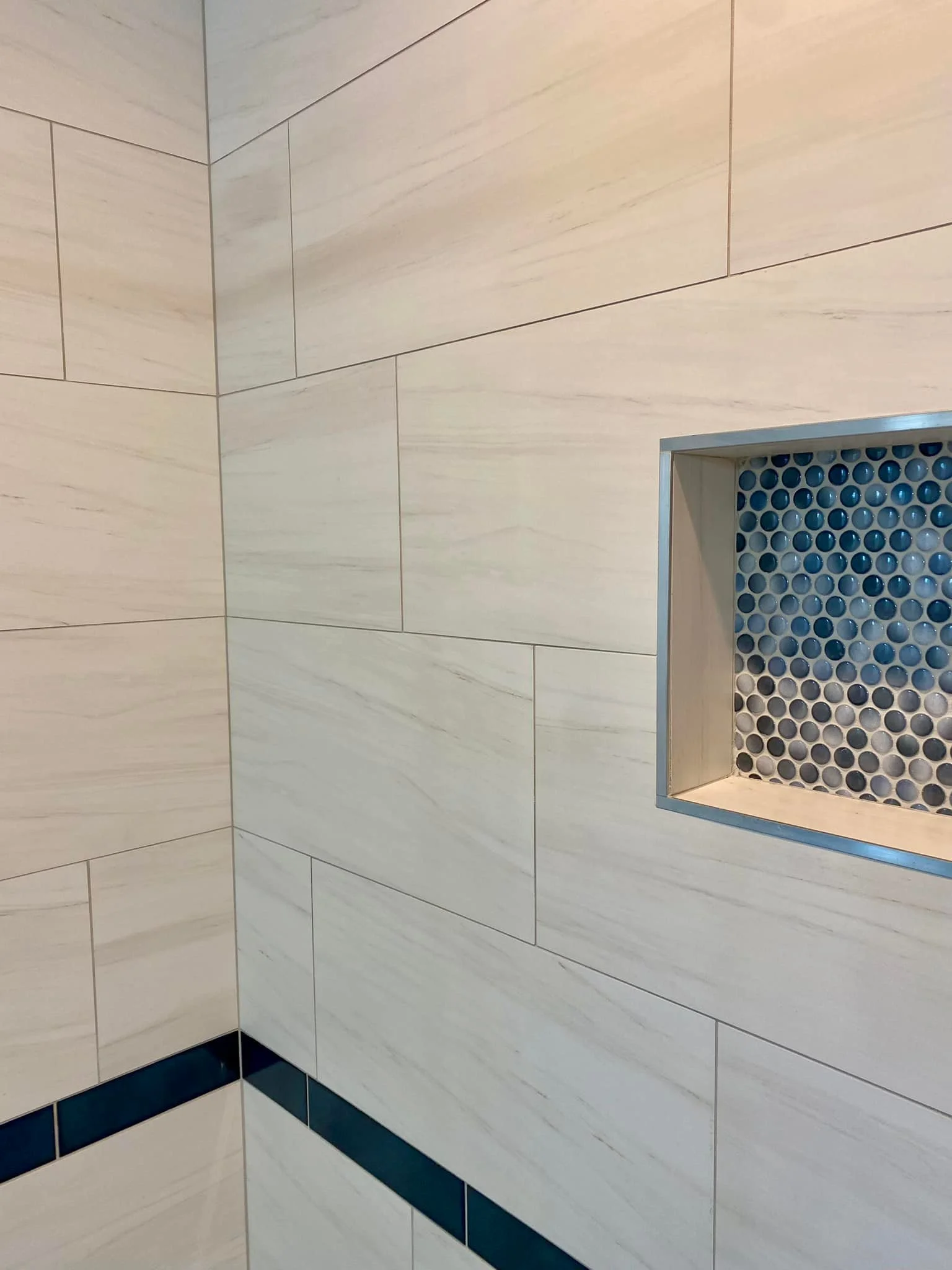 Close-up of a tiled shower wall with beige tiles, a blue accent strip, and a recessed shower niche lined with small blue and gray mosaic tiles.