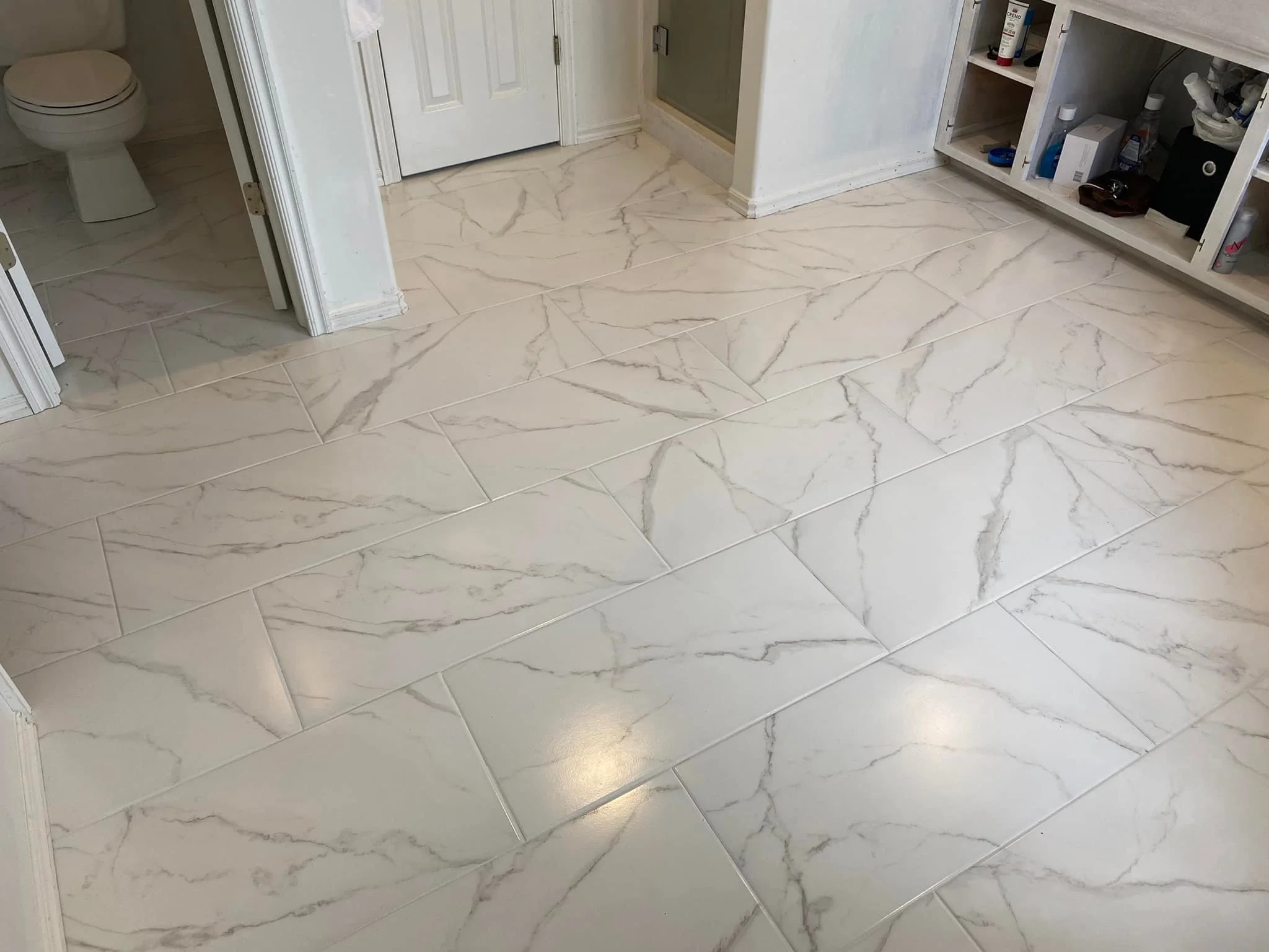 Bathroom with beige marble-look tile flooring, a toilet in a small room, and a white vanity with open shelves containing various toiletries and items.