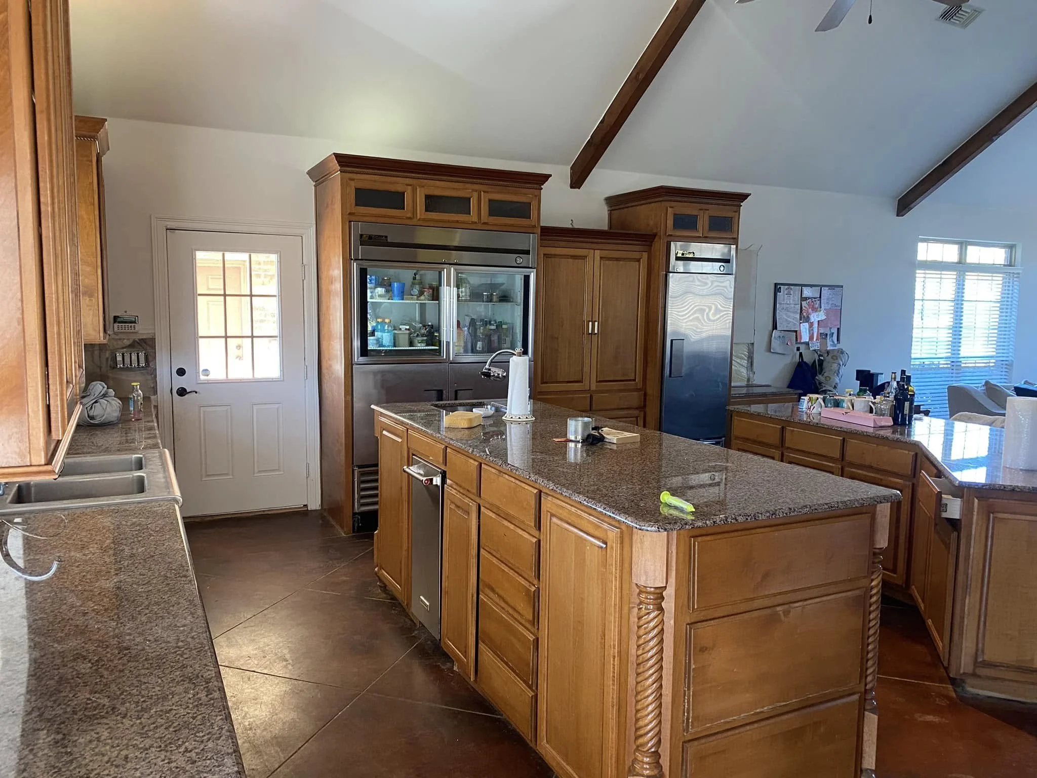Kitchen with wooden cabinets, granite countertops, stainless steel appliances, and a kitchen island. There are some items including paper towels, utensils, and bottles on the counters. A door leads outside, and there are windows with blinds in the background.