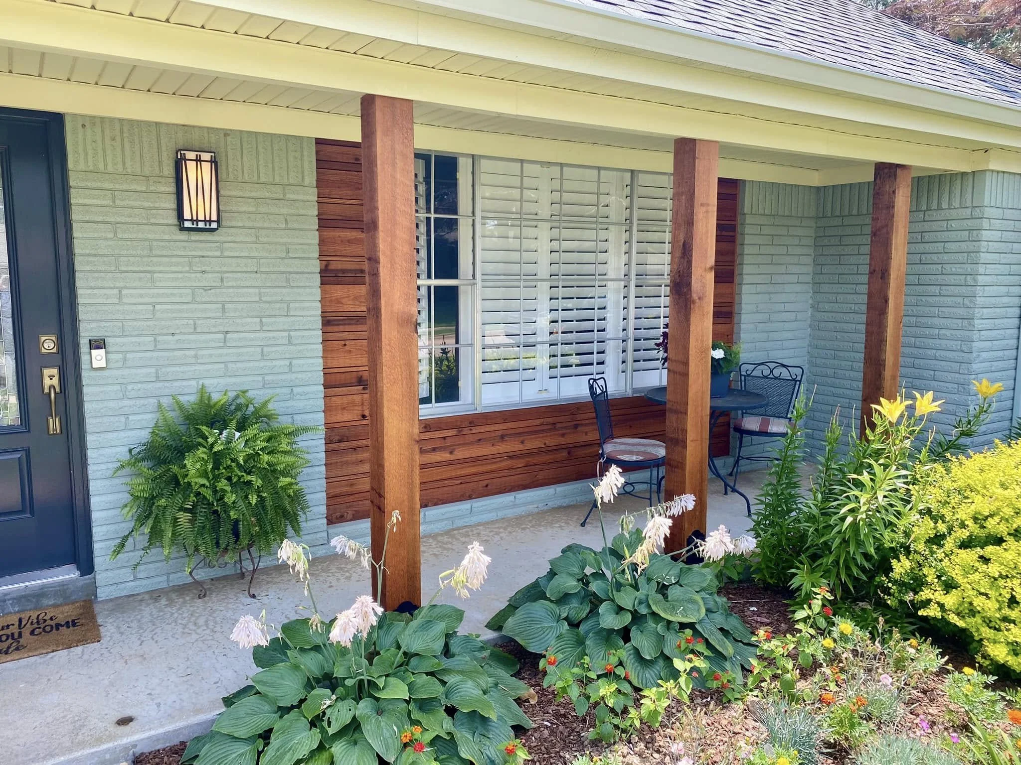 Front porch of a house with a blue brick wall, wooden pillars, a small table with two chairs, surrounded by green plants and colorful flowers.