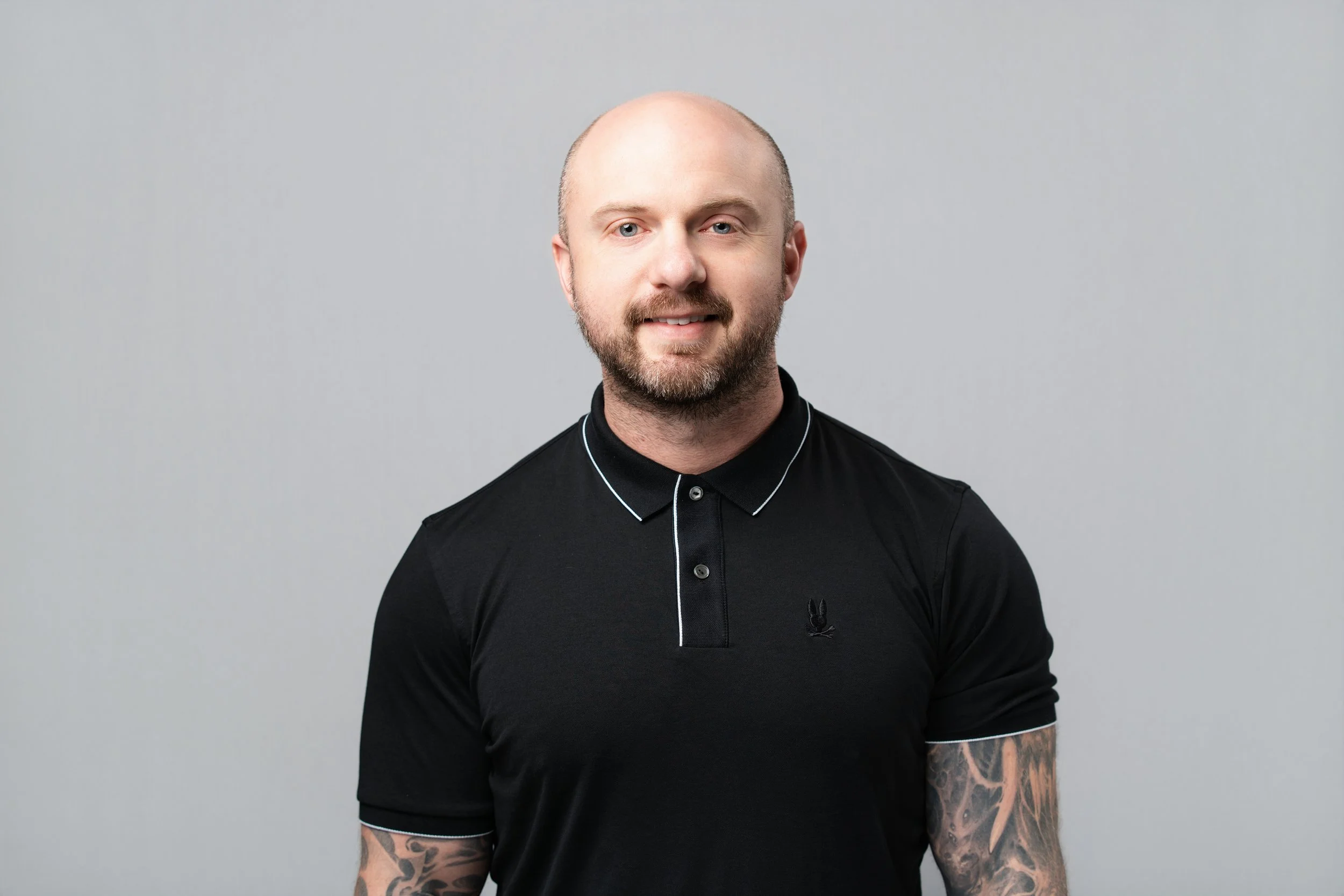 A portrait of a young man with a light complexion, and blue eyes, wearing a dark blue button-up shirt, smiling against a gray background. Taylor Dasch.
