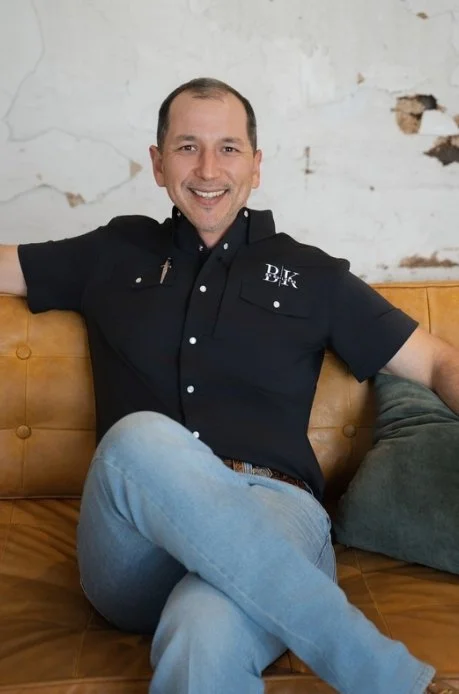 A man in a black polo shirt with a logo stands outdoors next to a wooden structure, smiling with his hands in his pockets. Background includes trees and a house.
