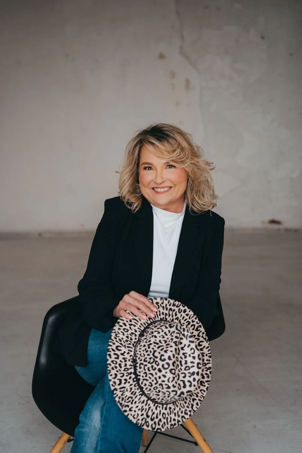 A smiling woman with wavy blonde hair wearing a white button-up shirt, sitting on a black chair in front of a plain gray wall.