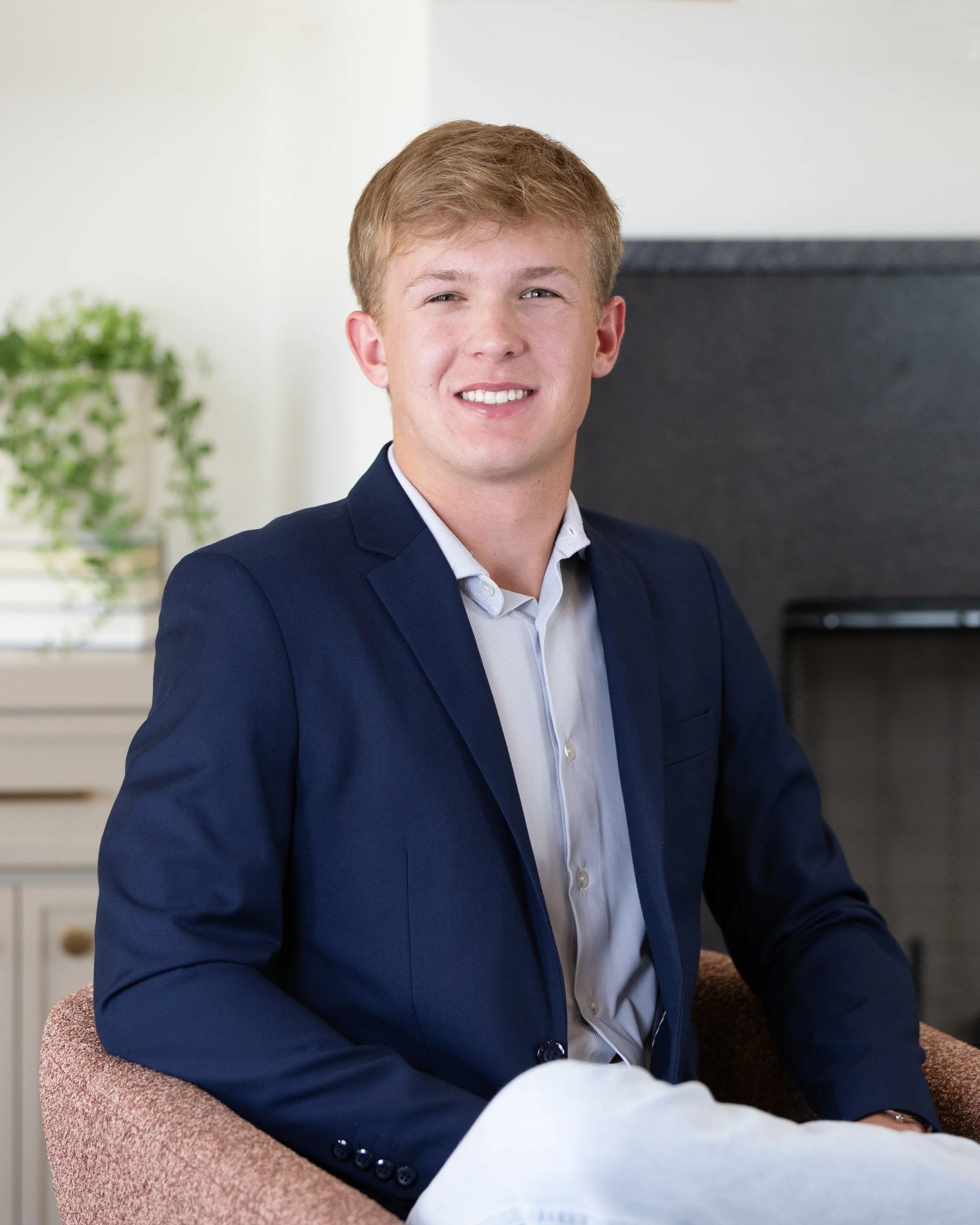 A young man with blonde hair wearing a dark shirt and blue suit jacket, smiling in front of a plain light-colored wall.