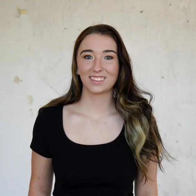 A young woman with long, wavy brown hair with blonde highlights, wearing a black short-sleeved top, smiling in front of a neutral, lightly textured wall.