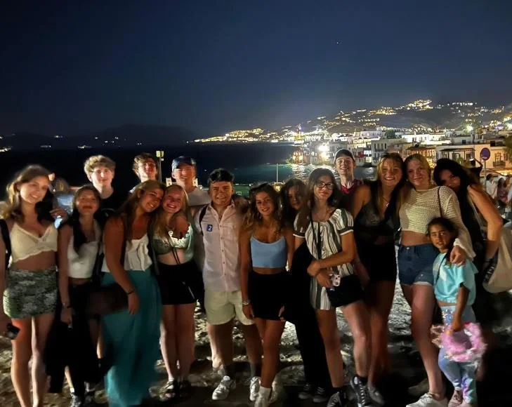 Group of young friends posing together on a beach at night, with a brightly lit hillside town in the background.