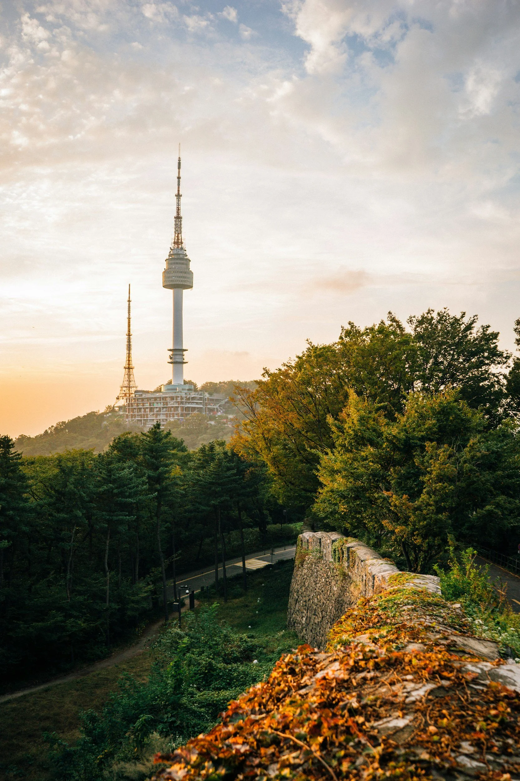 View of Namsan Seoul Tower on a hill at sunset, surrounded by trees and a stone wall in autumn.