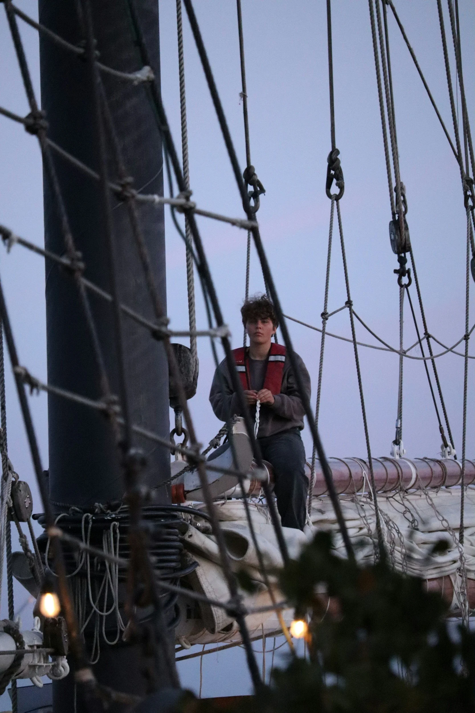 A young man sitting on the mast of a sailing ship, surrounded by rigging and ropes, during twilight or dusk.