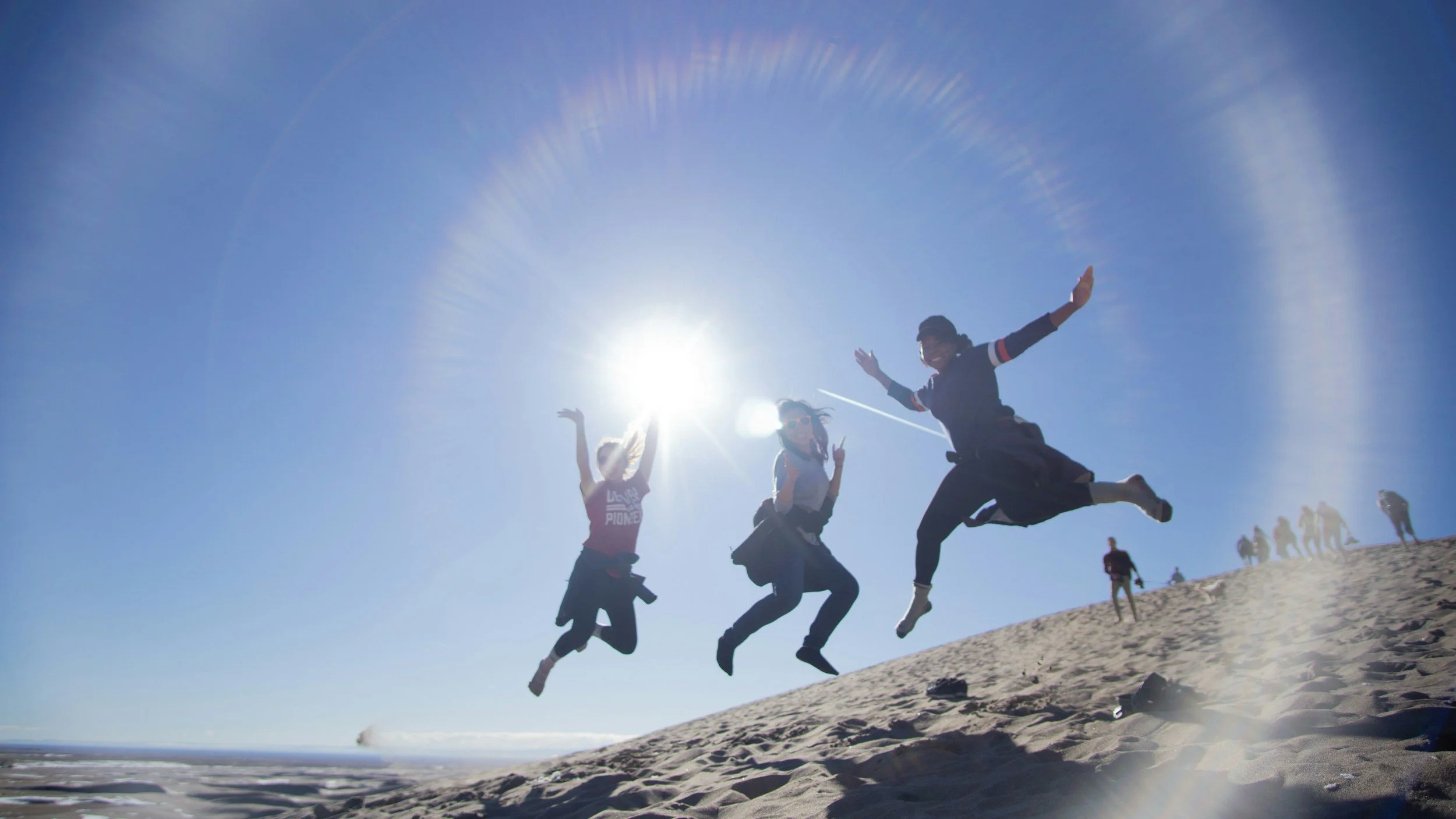 Three people jumping and smiling on a sandy beach with the sun shining brightly in the clear blue sky, a group of people in the background enjoying the day.