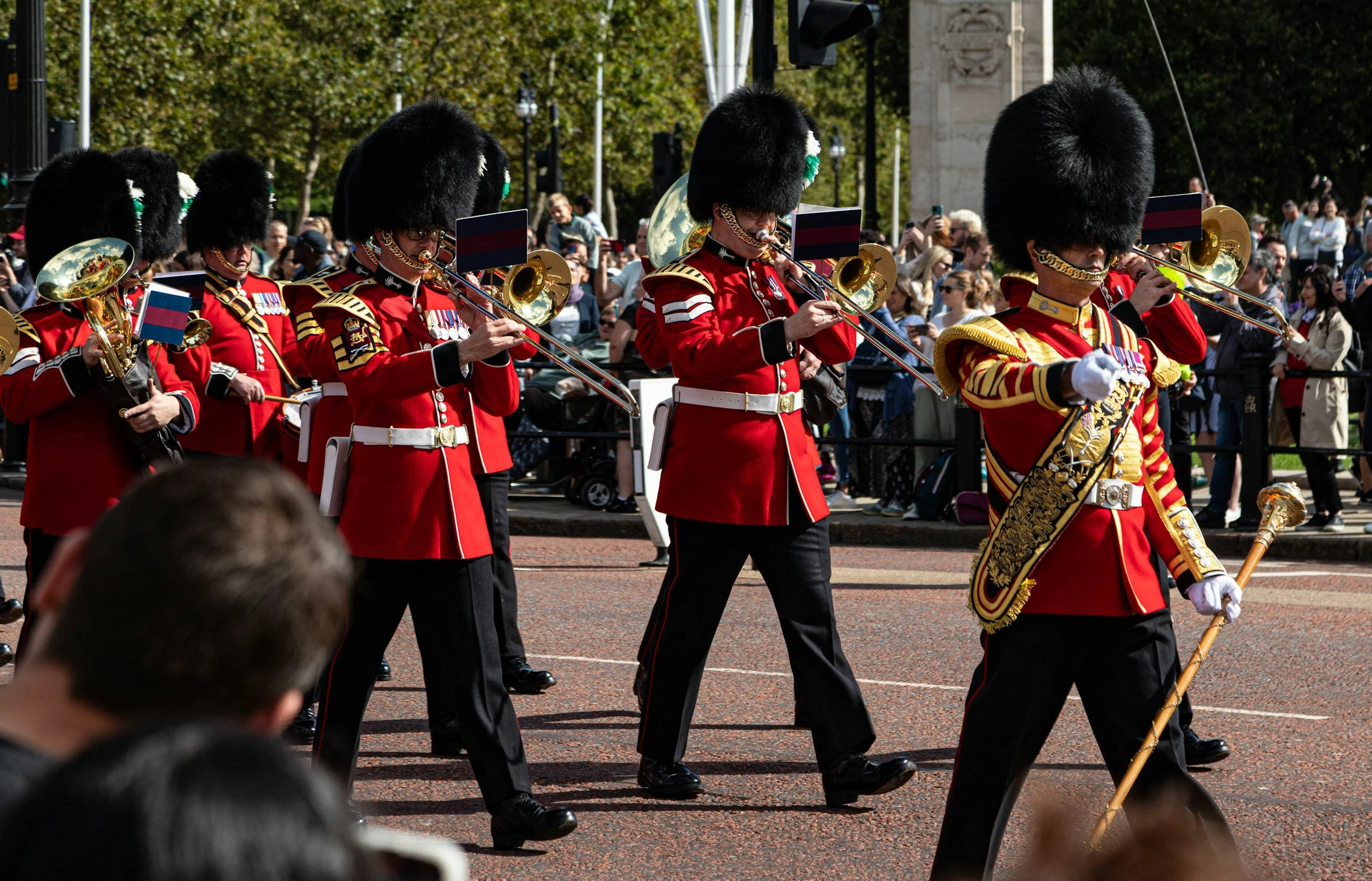 British soldiers in ceremonial uniforms marching in a parade, wearing red coats, black bearskin hats, and carrying musical instruments.