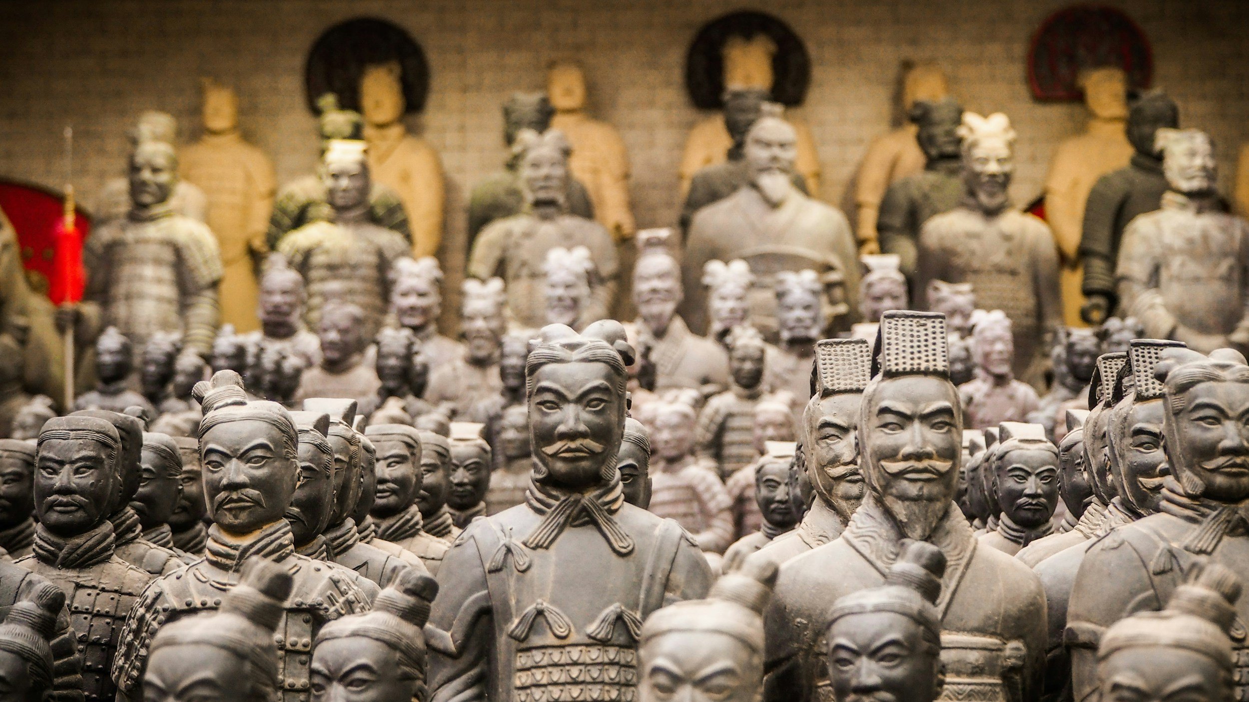 Collection of ancient Chinese terracotta warrior statues, with detailed facial features, armor, and traditional clothing, arranged in multiple rows in a museum exhibit.