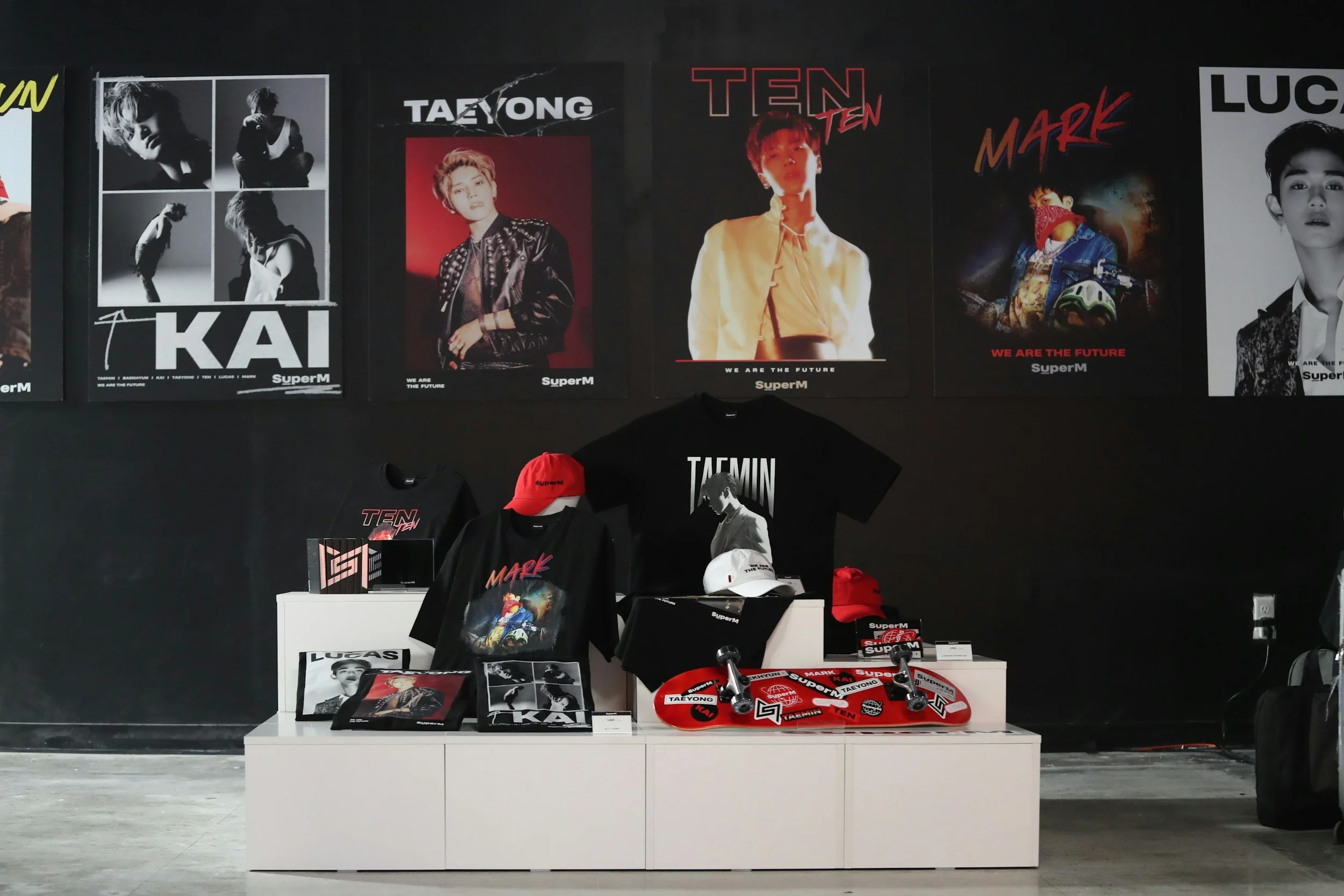 Display of K-pop merchandise featuring posters and apparel of Taemin, Mark, and Lucas, including t-shirts, caps, and skateboards in a retail setting.