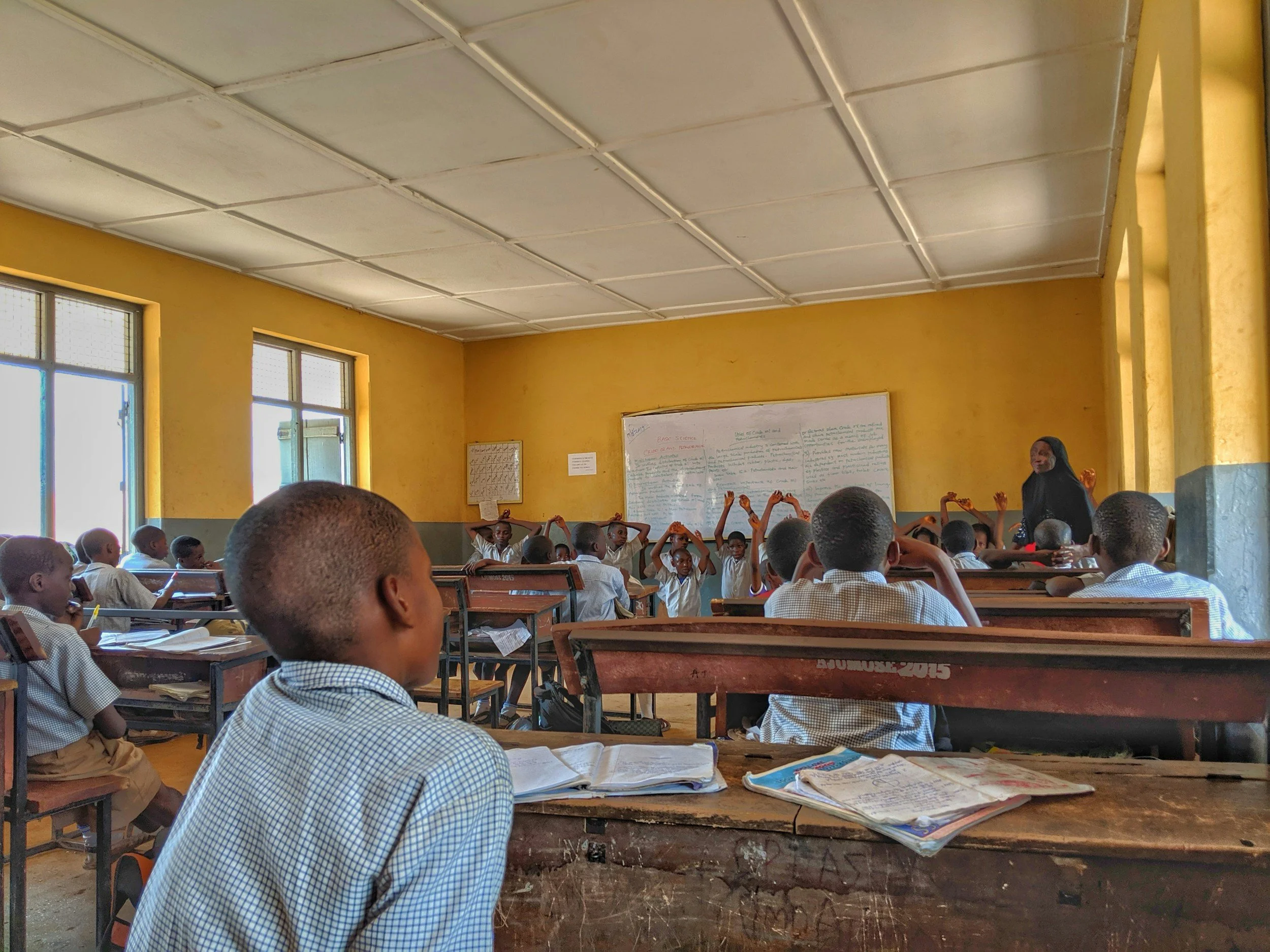 Classroom with students seated at wooden desks, some with notebooks, facing a teacher near the whiteboard. Students in white uniforms, yellow walls, and large windows letting in natural light.