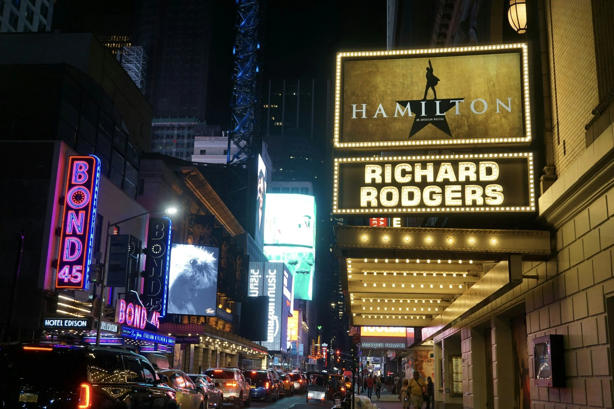 Nighttime scene of Times Square with bright illuminated signs, billboards, and marquees, including Hamilton and Richard Rodgers theater signs, with cars and pedestrians on the street.