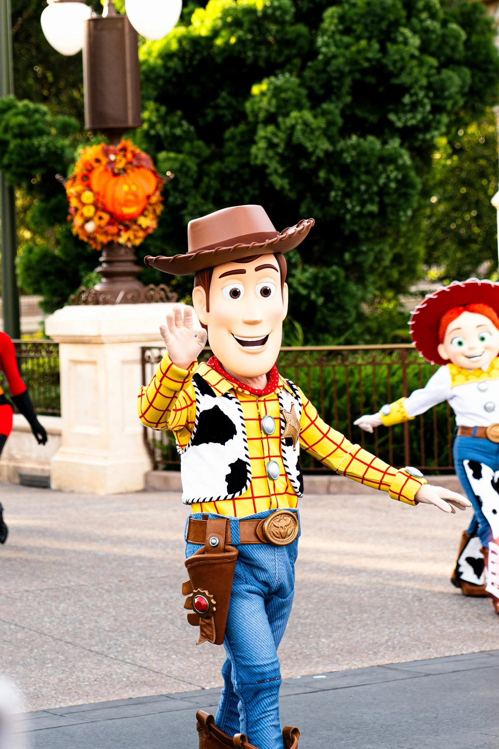Costumed characters of Woody and Jessie from Toy Story waving while standing outdoors at a theme park, with Halloween decorations in the background.
