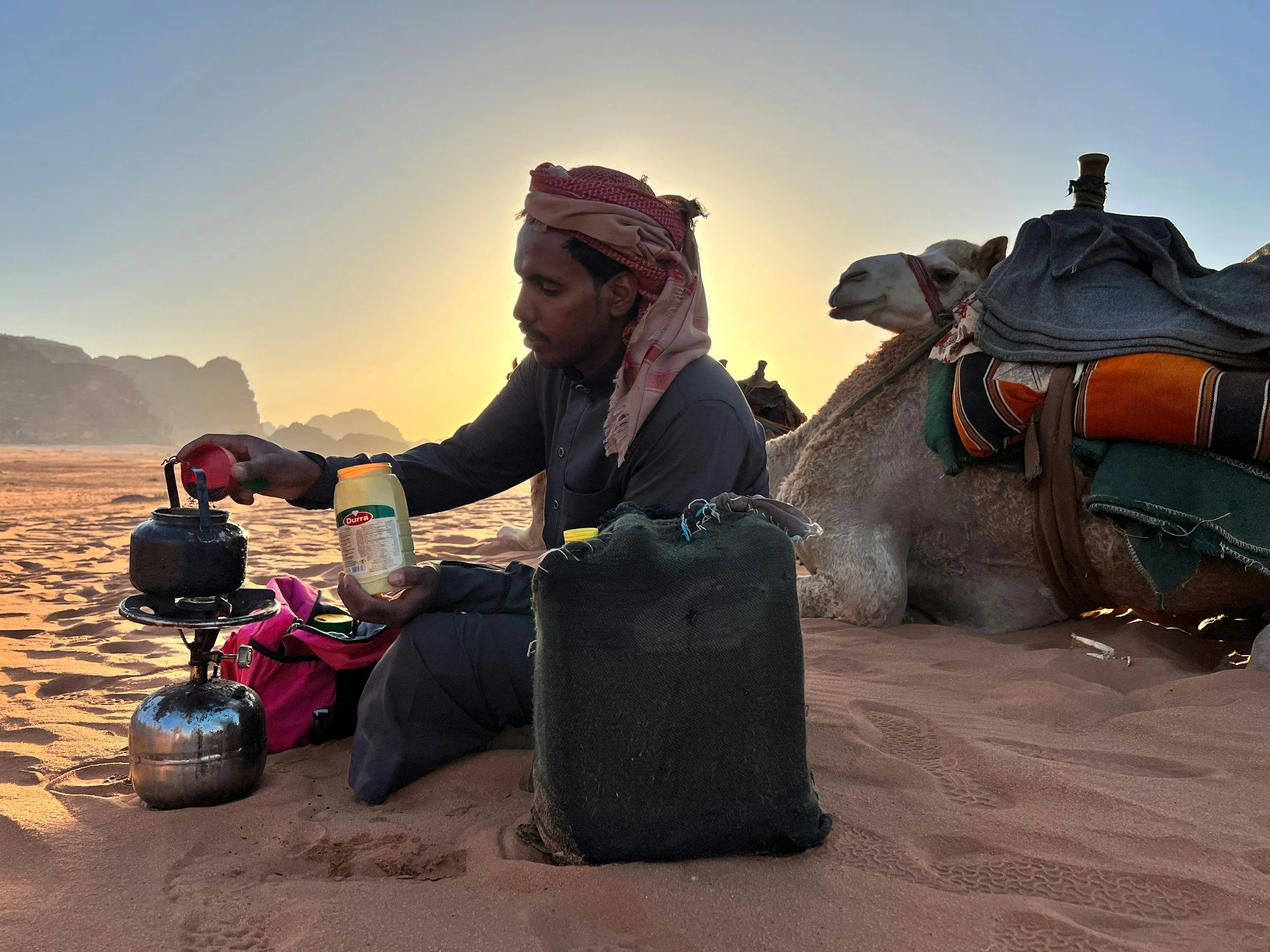 nomad pouring teas in desert with a bright blue sky, with a friendly looking camel lying beside the camp area.