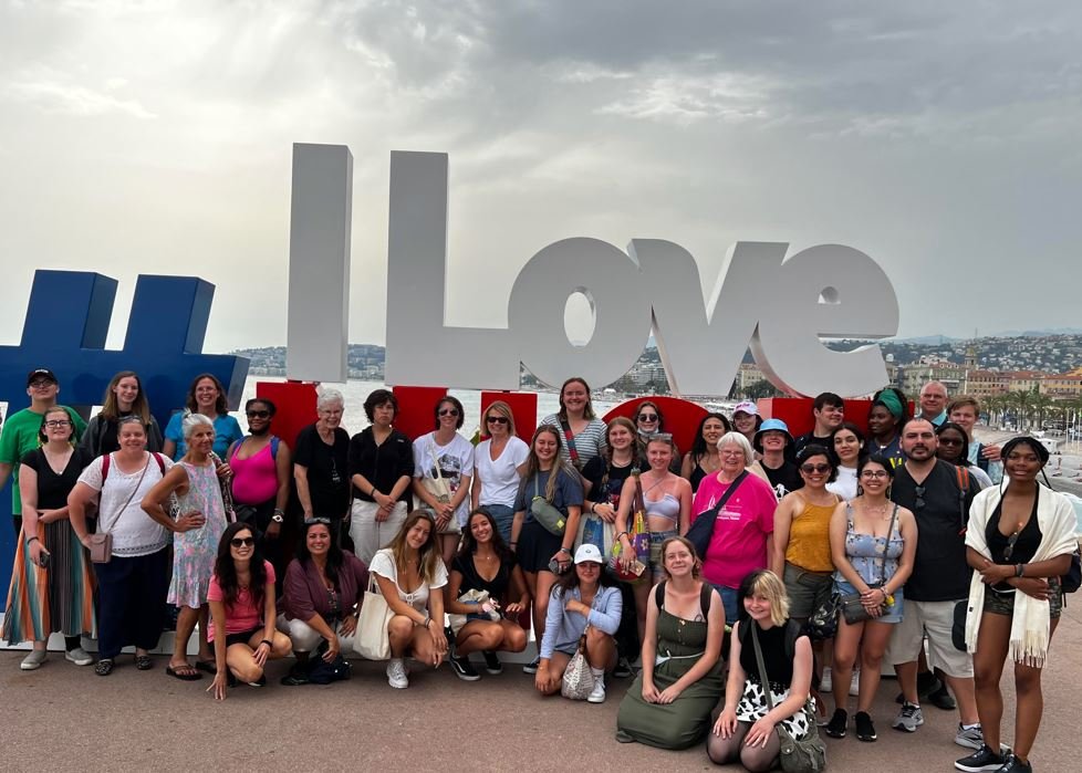 A group of diverse people posing in front of large white and blue letters spelling 'I Love' at a waterfront location with cityscape in the background.