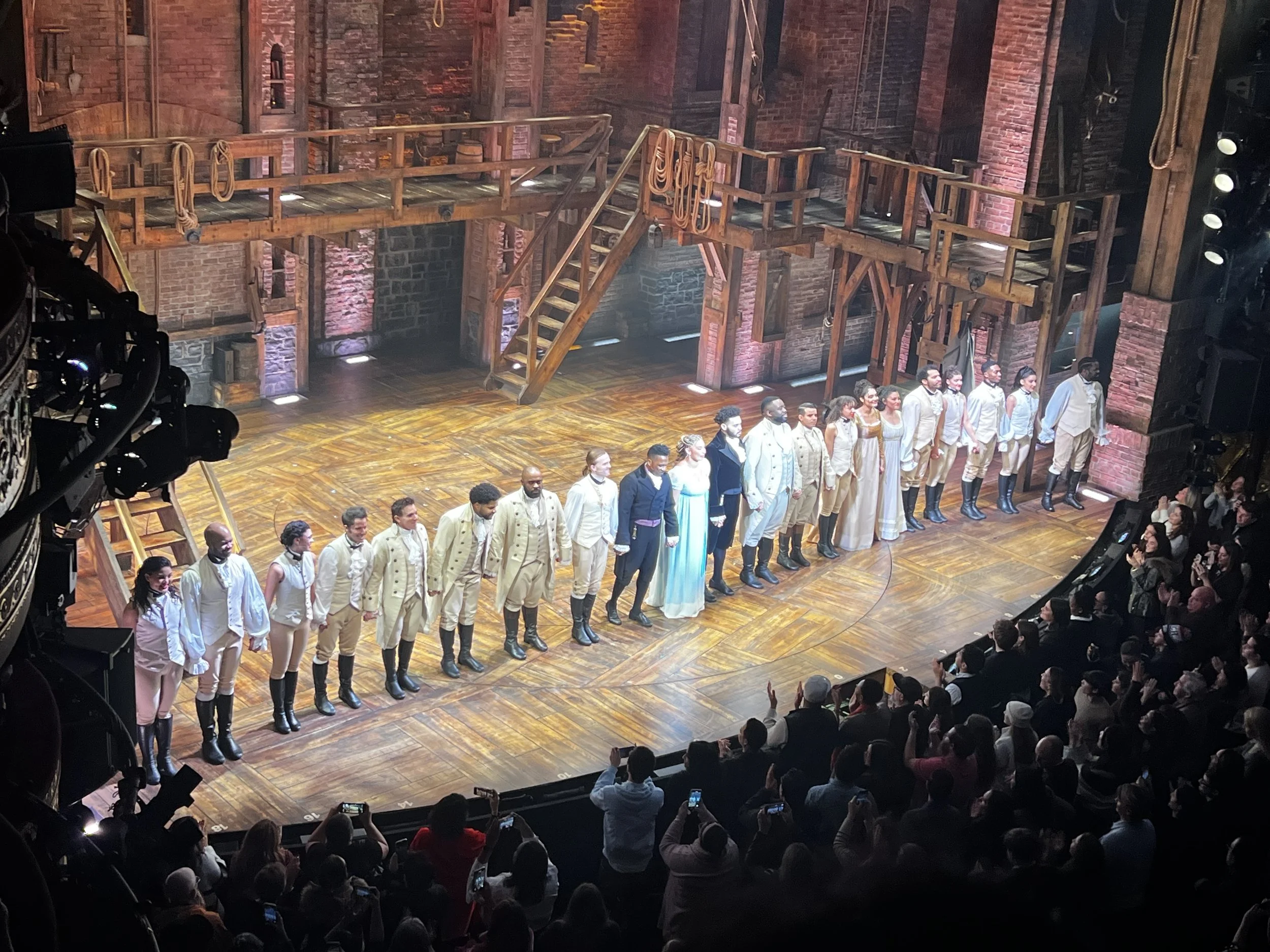 Cast members of a theater show take a bow on stage at the end of a performance, with an audience clapping and taking photos.