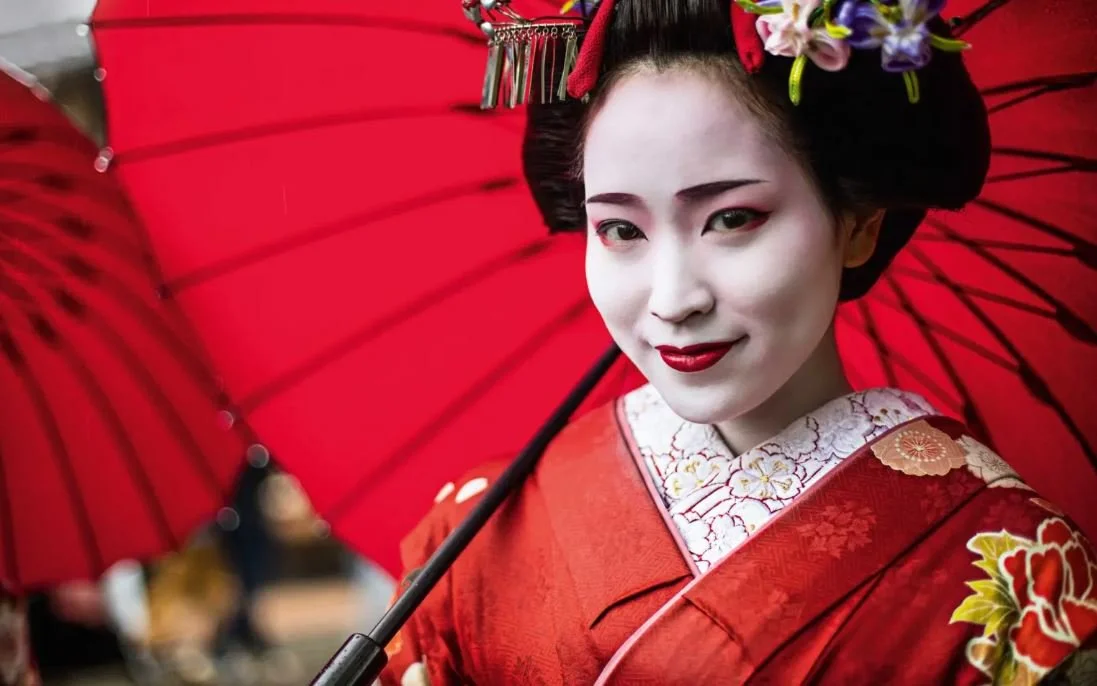 A woman dressed as a Japanese geisha, holding a traditional red umbrella, with a red background, wearing a red kimono with floral patterns, and makeup with white face paint and red lipstick.