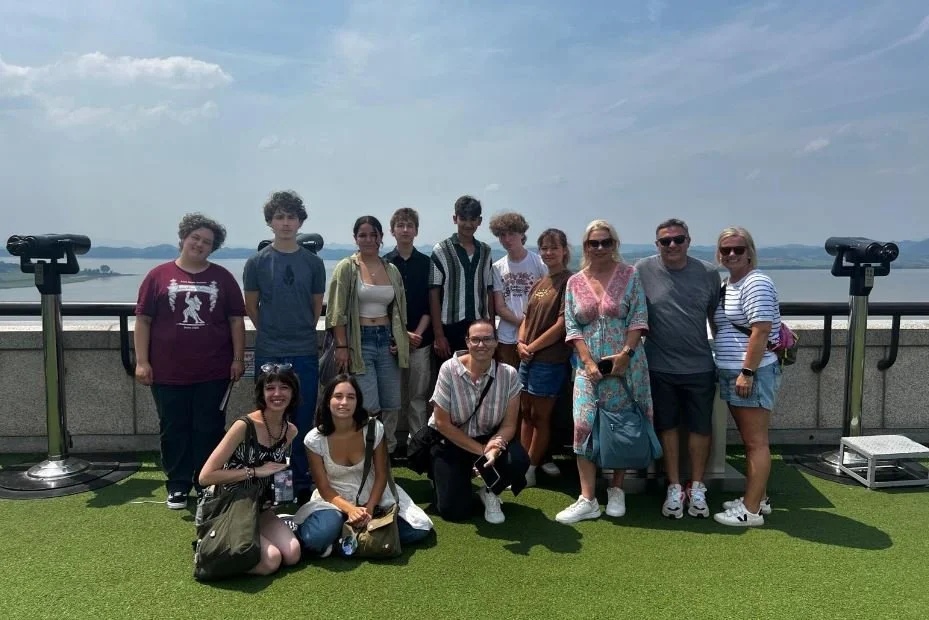 Group of people standing on a rooftop with telescopes, overlooking a body of water and distant mountains under a partly cloudy sky.