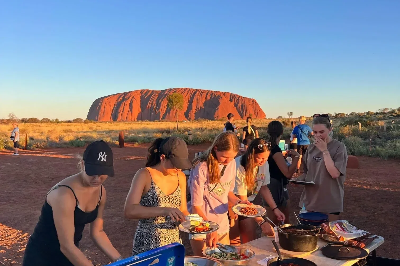 Tourists enjoying a meal outdoors in front of Uluru at sunset, with the iconic large red rock in the background.