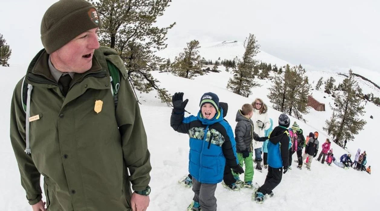 A park ranger with a badge on his green uniform stands in front of a line of children and adults in a snowy landscape, some of whom are smiling and waving.