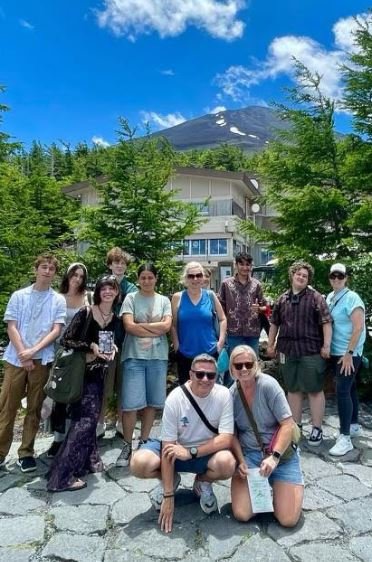 Group of ten people posing outdoors in front of a mountain and trees, with a building in the background.