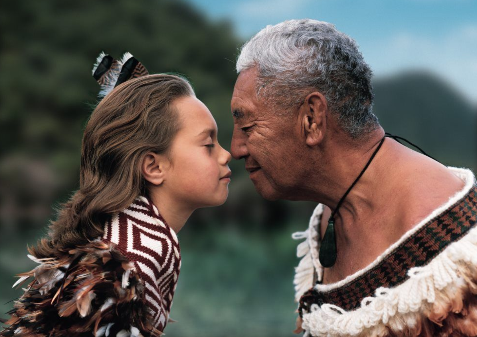 A young girl and an older man face each other with foreheads touching outdoors, both with closed eyes, wearing traditional tribal attire, with a green mountainous landscape in the background.