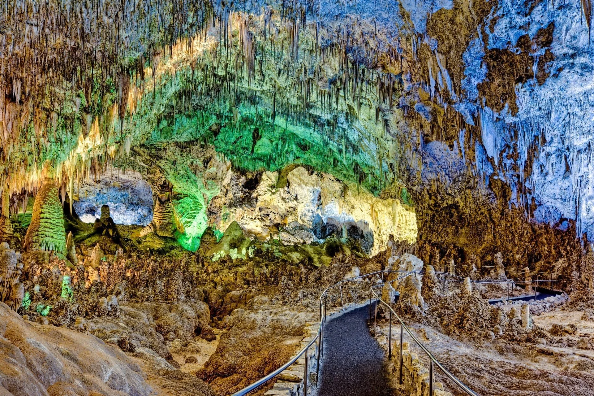 A colorful cave interior with stalactites and stalagmites, illuminated with multicolored lights and a pathway with railing for visitors.