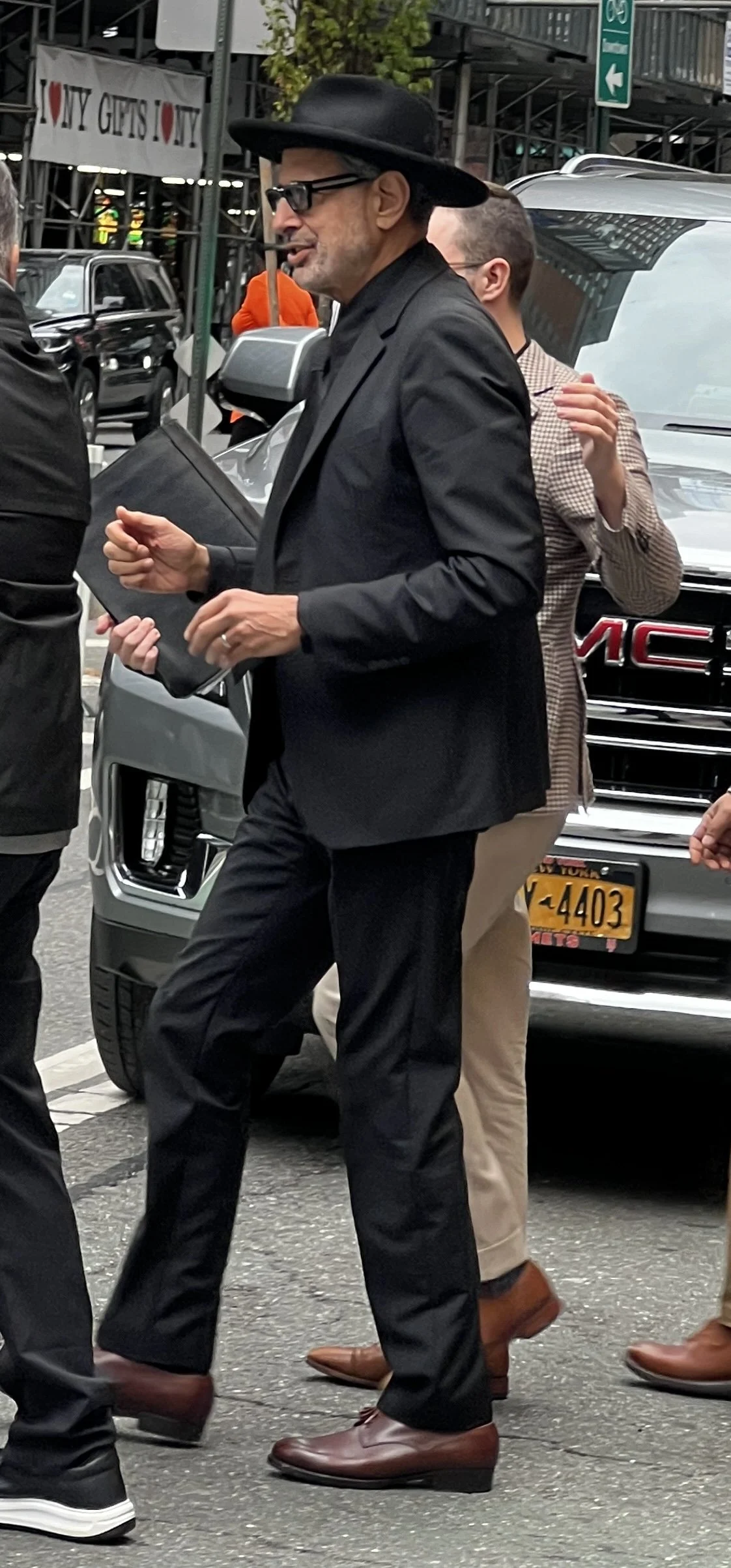 A man wearing a black hat, glasses, and a black suit holding a black tablet or folder, walking on a city street with cars and pedestrians around.