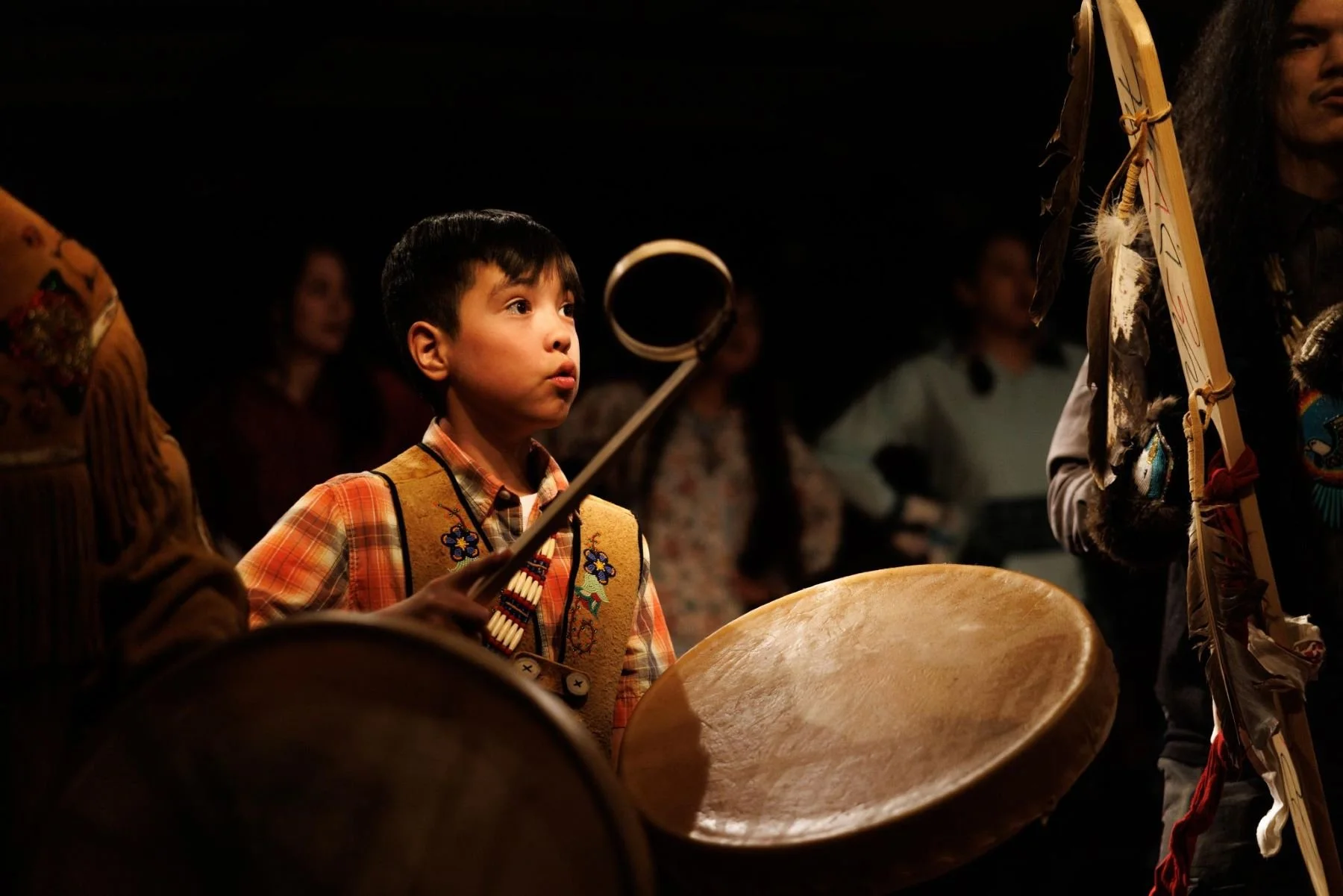 A young boy playing a drum during a cultural performance, surrounded by other performers in traditional attire.