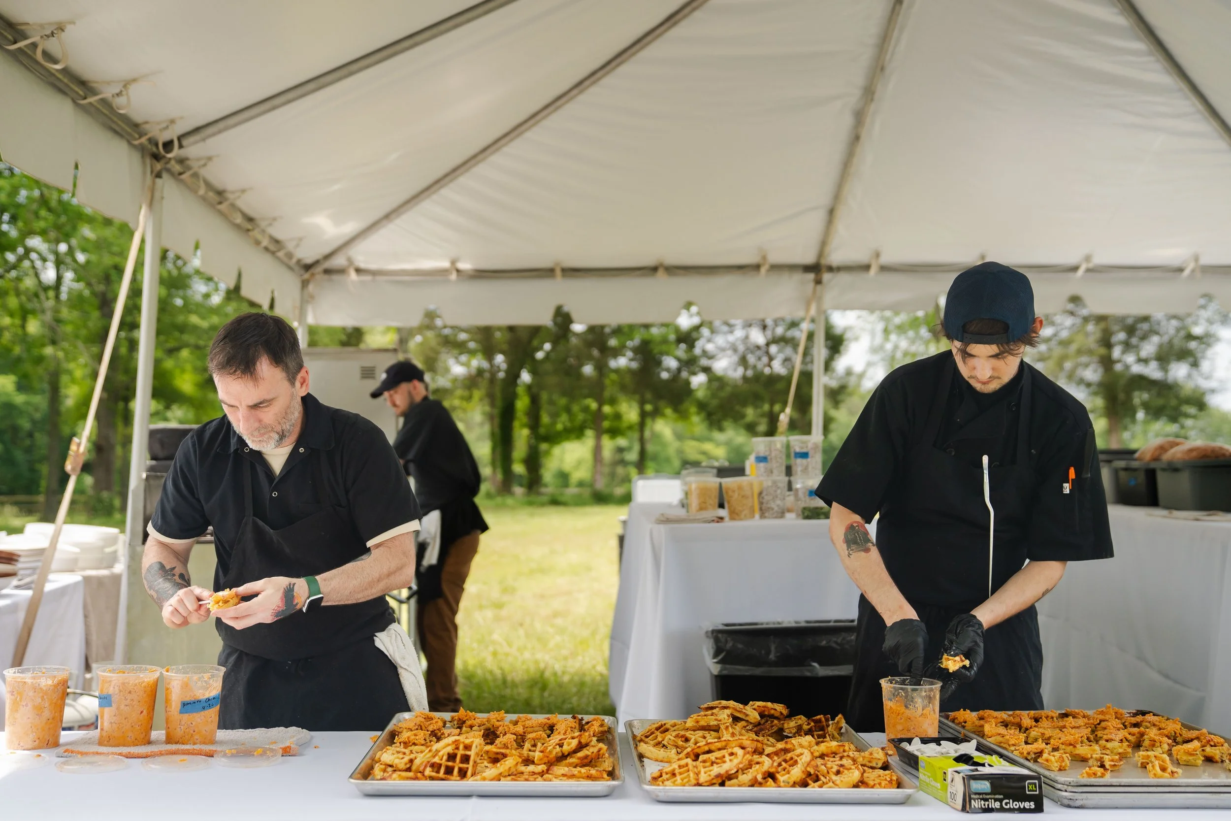 Three men working at an outdoor food event, preparing and serving waffle sandwiches under a white tent, with a table of ingredients and food in front of them, against a background of trees.