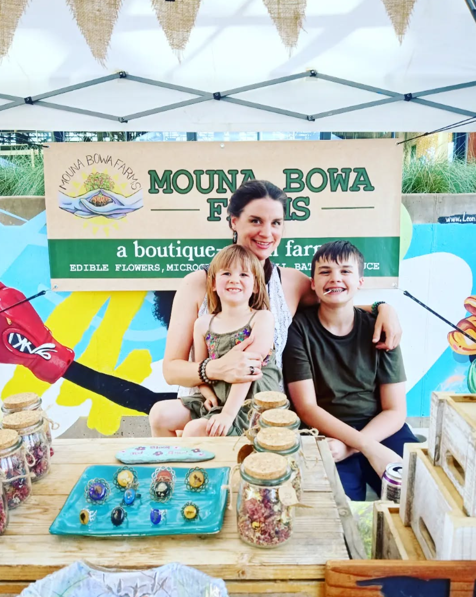 A woman with two children smiling at a booth for Mouna Bowa Fowers, which sells edible flowers, microgreens, and plants. The booth has jars of dried flowers and a blue tray with decorative rings. There is a colorful mural behind them and a white canopy above.