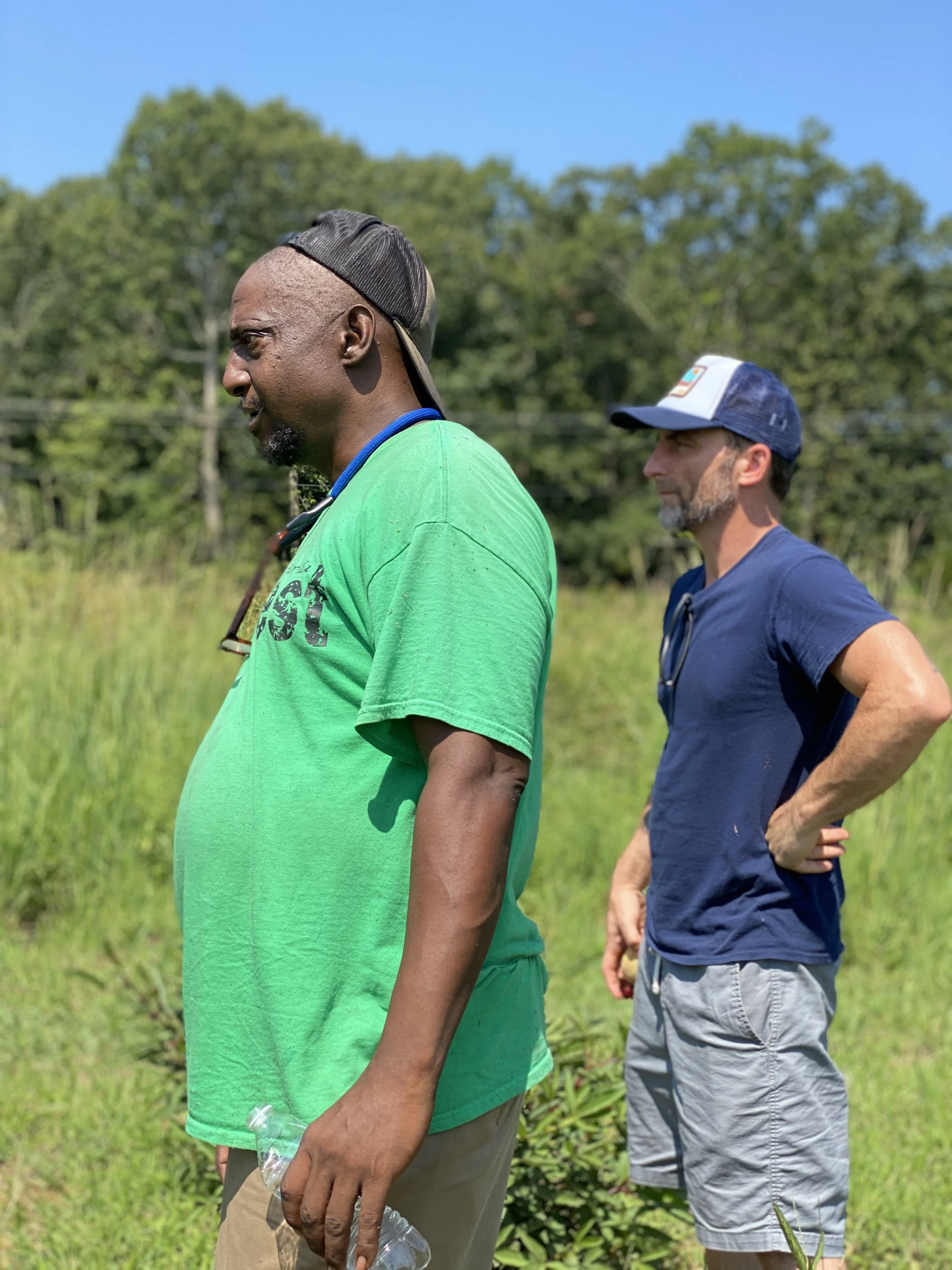 Two men standing outdoors in a grassy area with trees in the background, one in a green t-shirt and the other in a blue shirt and cap, looking to the side.