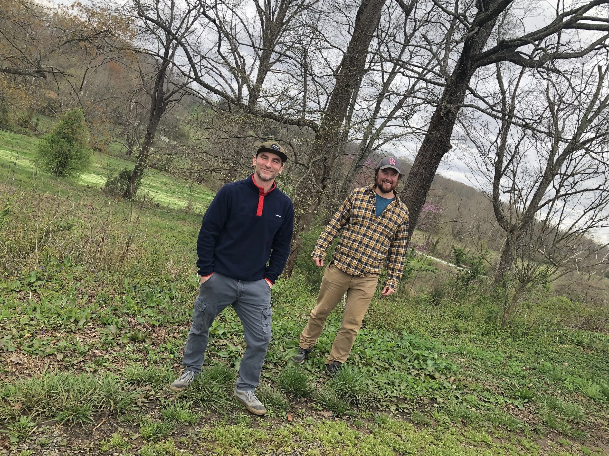 Two men standing outdoors in a grassy, tree-filled landscape on a cloudy day.
