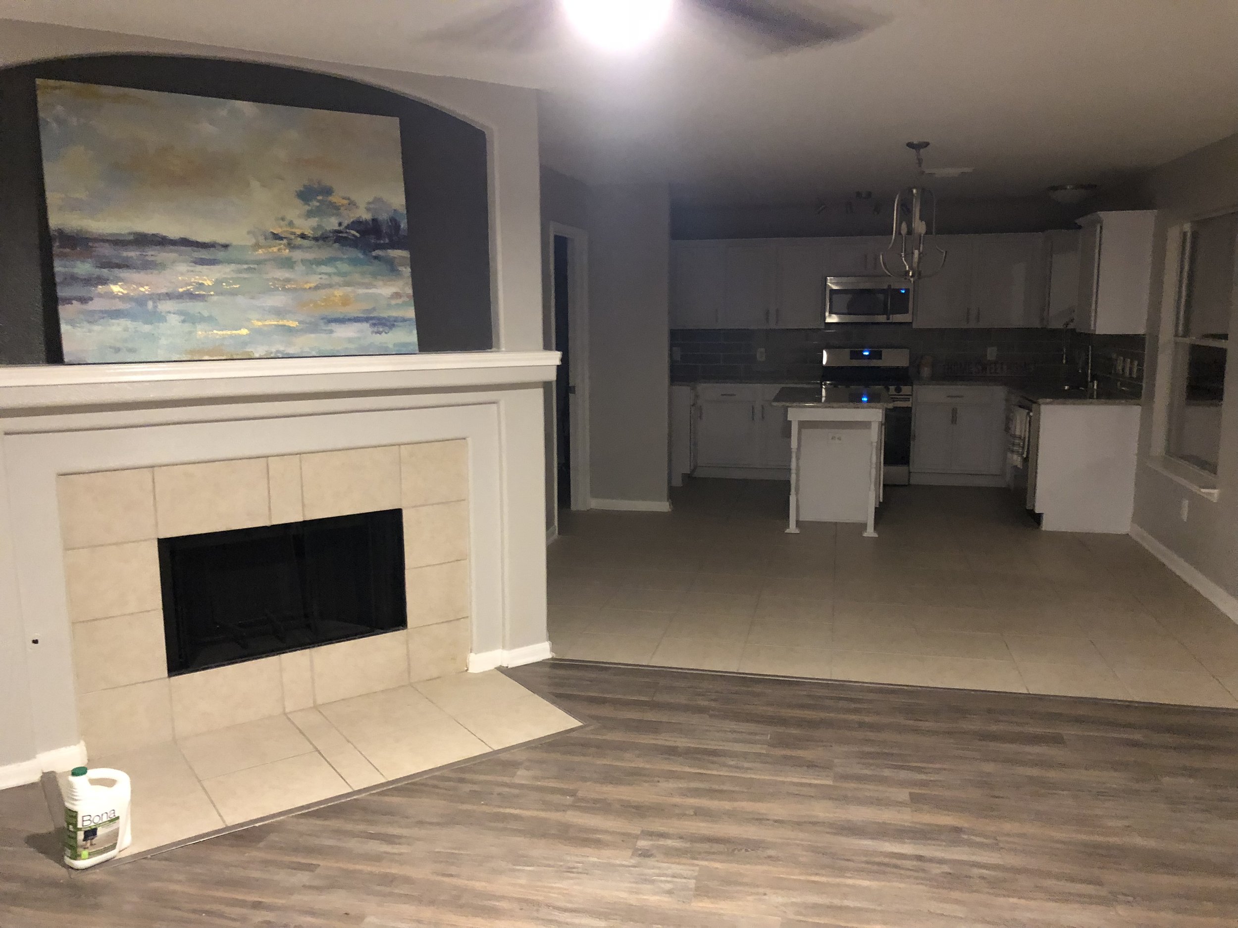 Living room with tiled fireplace and landscape painting, view of modern kitchen with stainless steel appliances and a small island, wooden and tiled flooring.