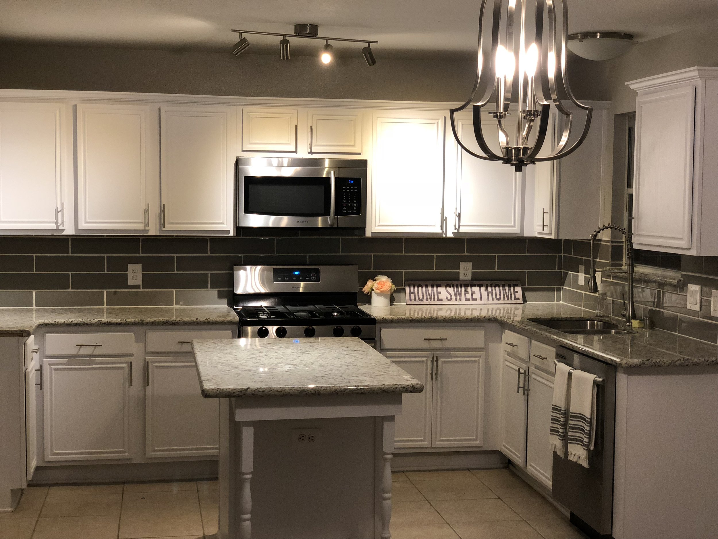 Modern kitchen with white cabinets, stainless steel appliances, a gray tile backsplash, granite countertops, and a central island. A chandelier and track lighting illuminate the space. A decorative sign reads "Home Sweet Home."
