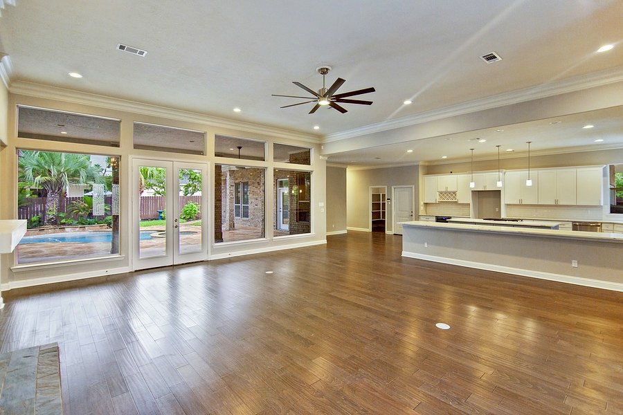 Spacious empty living room with wooden floors, large windows, and a view of the backyard pool. Open concept kitchen with modern design and recessed lighting. Ceiling fan in the center.