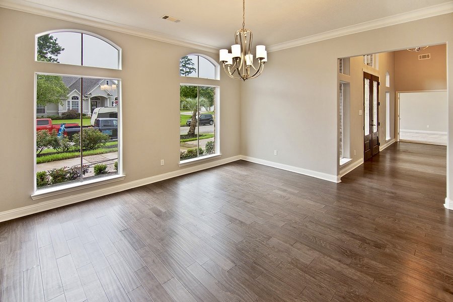 Empty living room with hardwood flooring, large windows, chandelier, and arched entryway, leading to a hall with doors and additional rooms.