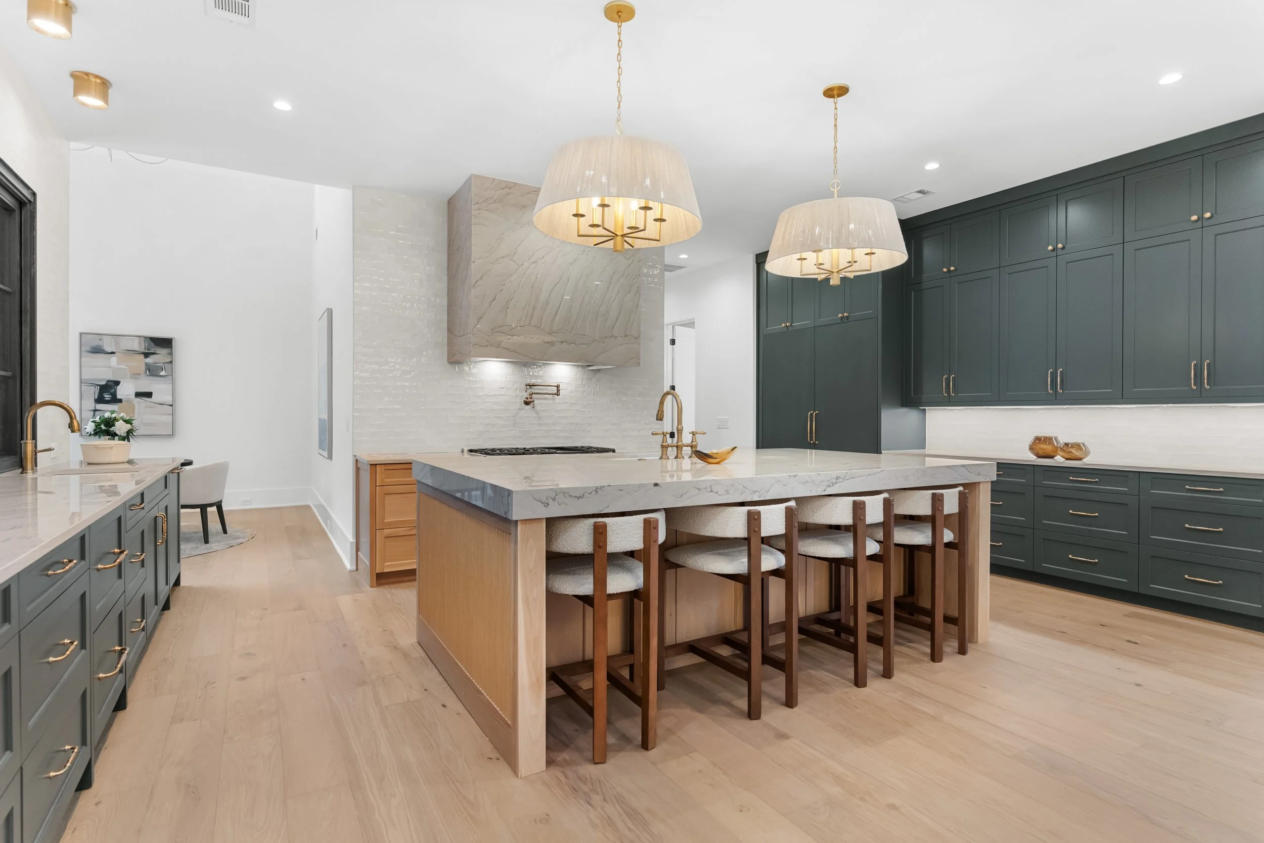 Modern kitchen with green cabinetry, a marble island with seating, and two pendant lights hanging above the island. Light wood flooring, white walls, and minimal decor.