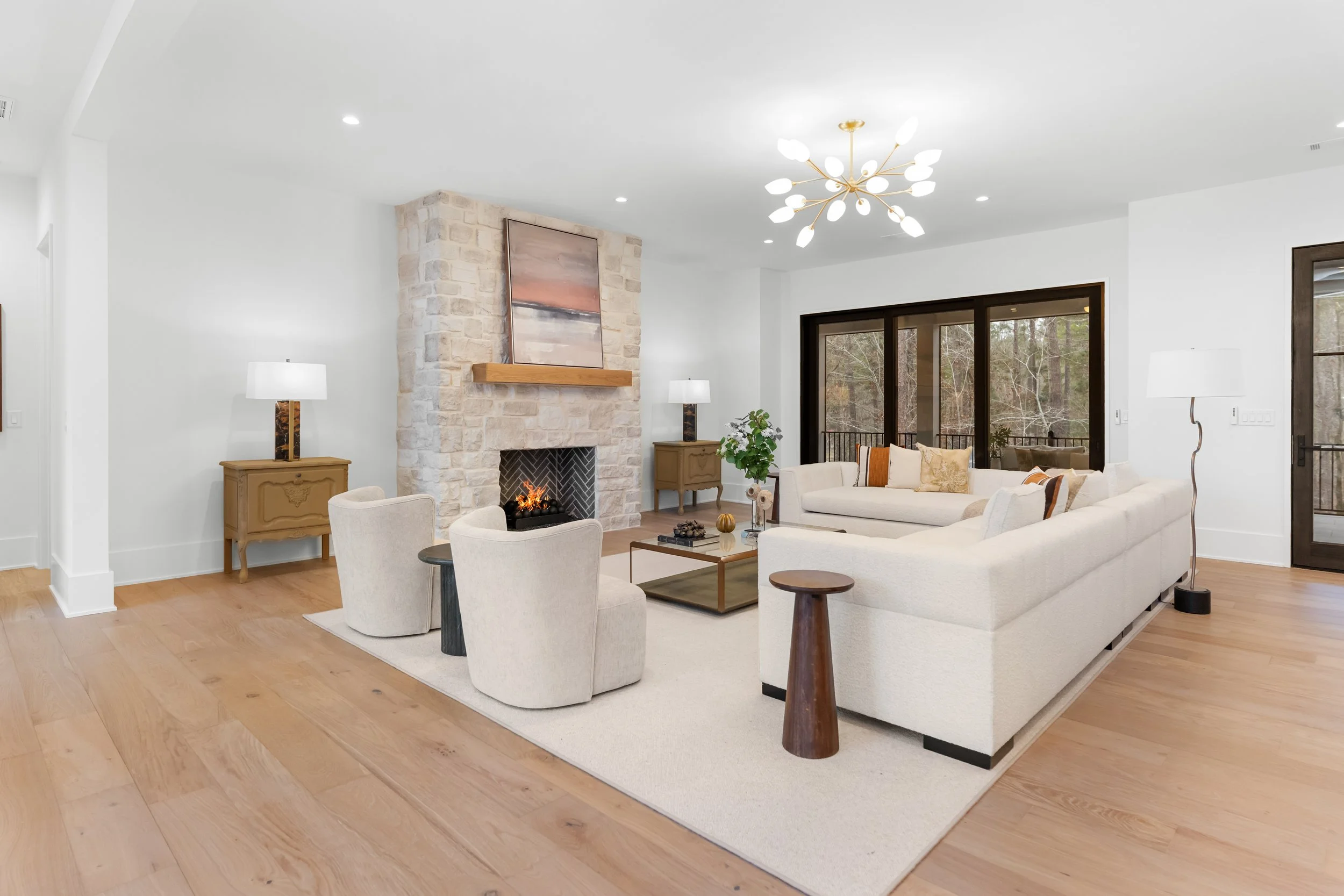 Living room with white walls, light wood flooring, a stone fireplace with a painting above, modern chandelier, two beige armchairs, a beige sectional sofa, side tables with lamps, and large sliding glass doors leading to a balcony with trees outside.