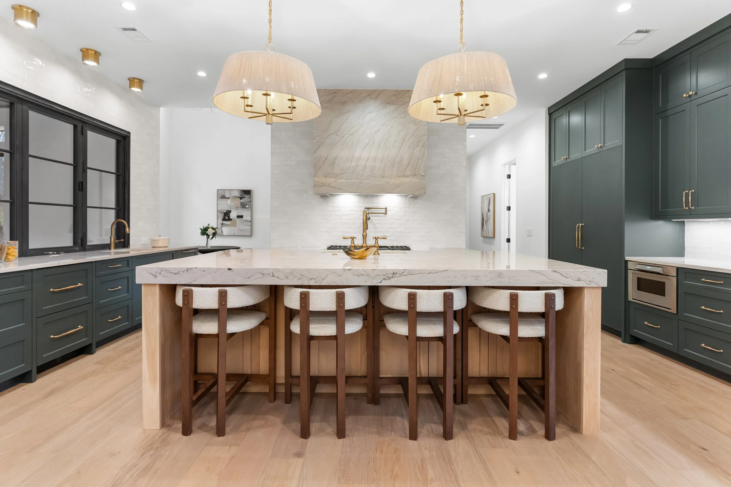 Modern kitchen with dark green cabinets, a large island with a marble countertop, beige bar stools, and gold accents, pendant lights, and hardwood flooring.