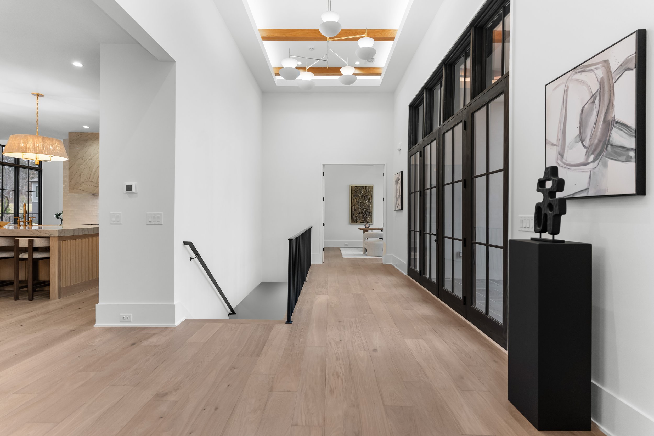 Modern interior hallway with light wood flooring, white walls, large black-framed glass doors on right, abstract artwork, black sculpture on pedestal, and staircase with black railing leading downstairs