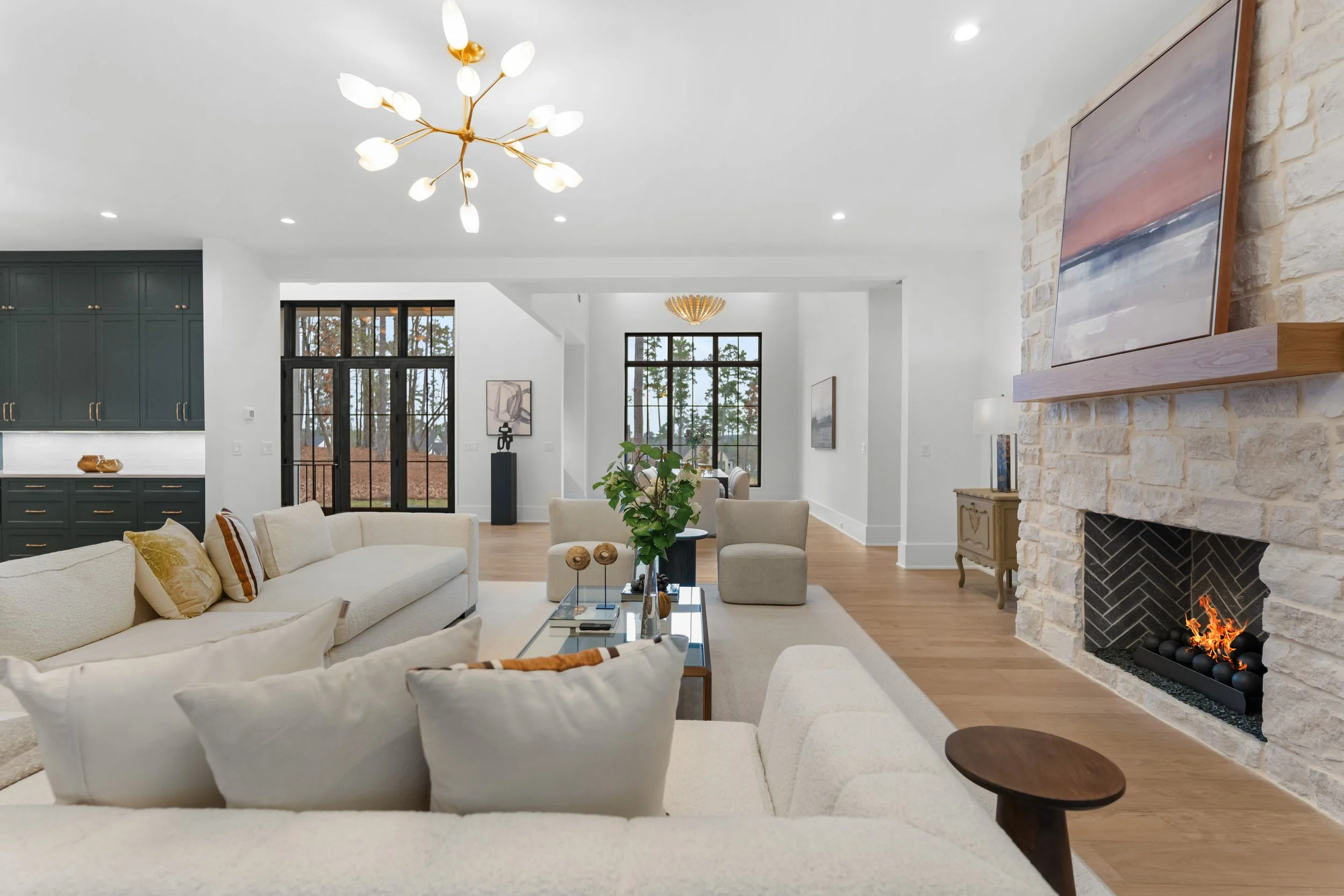 Modern living room with cream-colored sofas, a glass coffee table, a stone fireplace with a painting above, hardwood floors, and large windows showing trees outside.