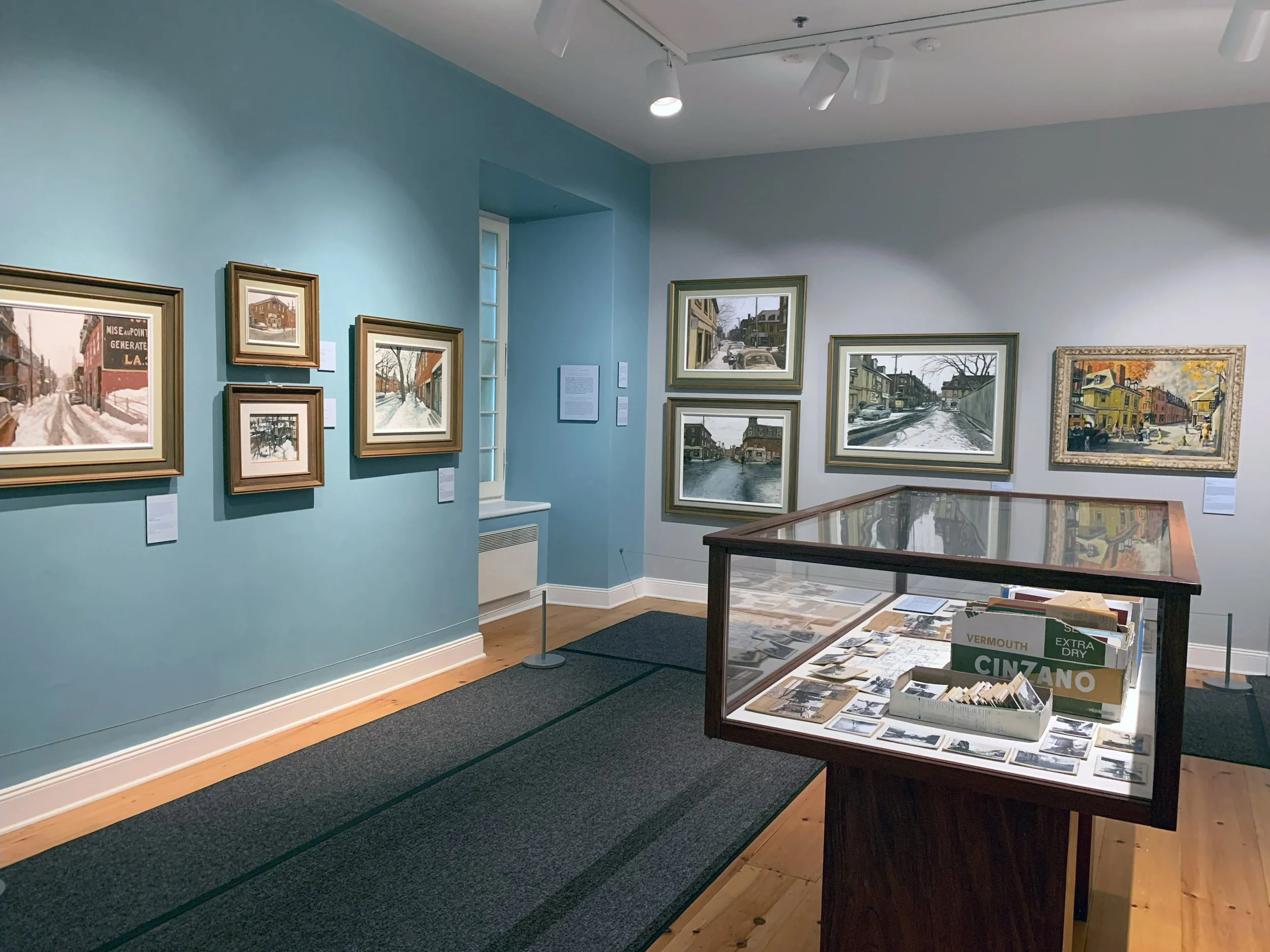 Art gallery showing framed landscape paintings on light blue and white walls, with a display case containing photographs and printed materials block to the right.