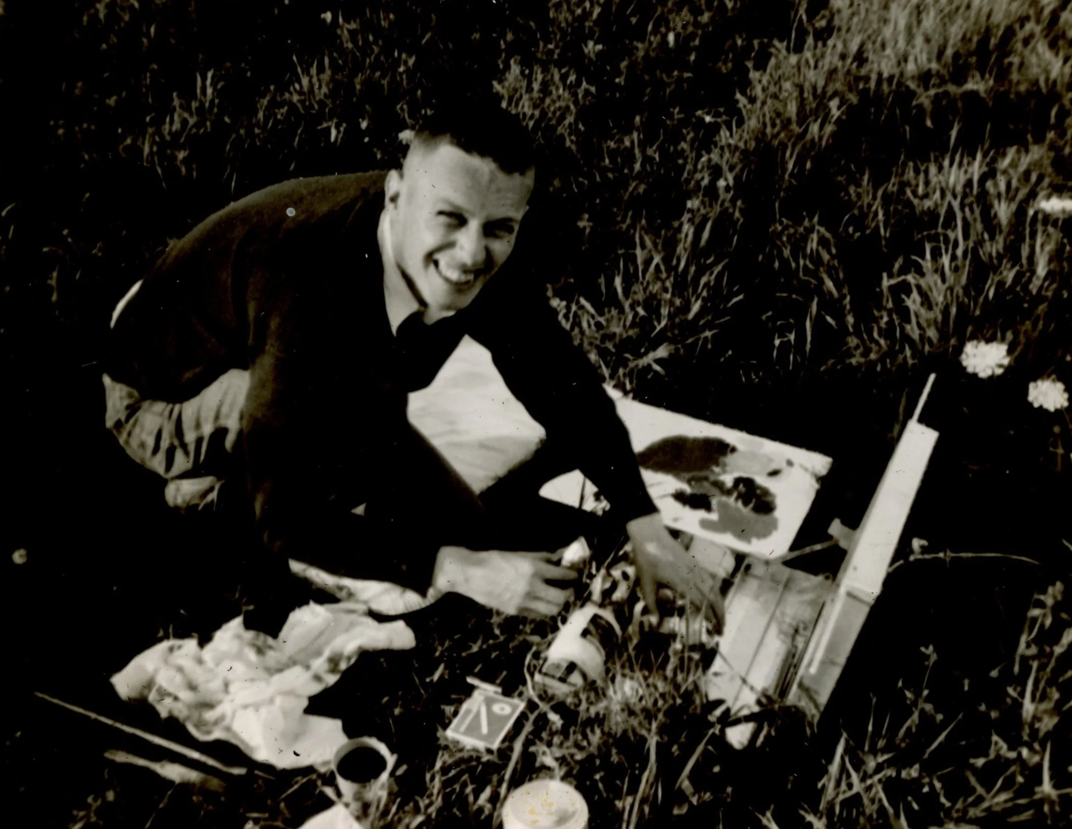 A person smiling and crouching in a field next to a model airplane, with a paint palette and brushes nearby.