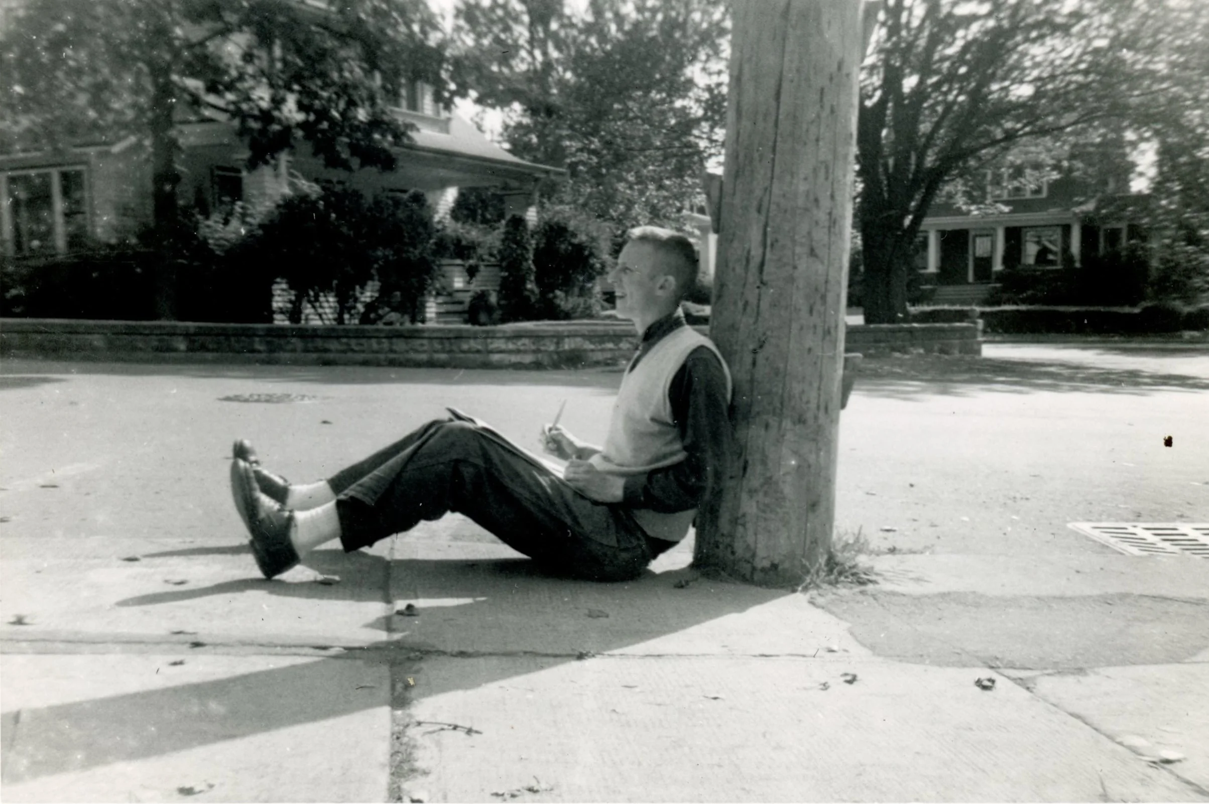 A young man sitting on a sidewalk, leaning against a utility pole, holding a notebook and pen, in a residential neighborhood with trees and houses in the background.
