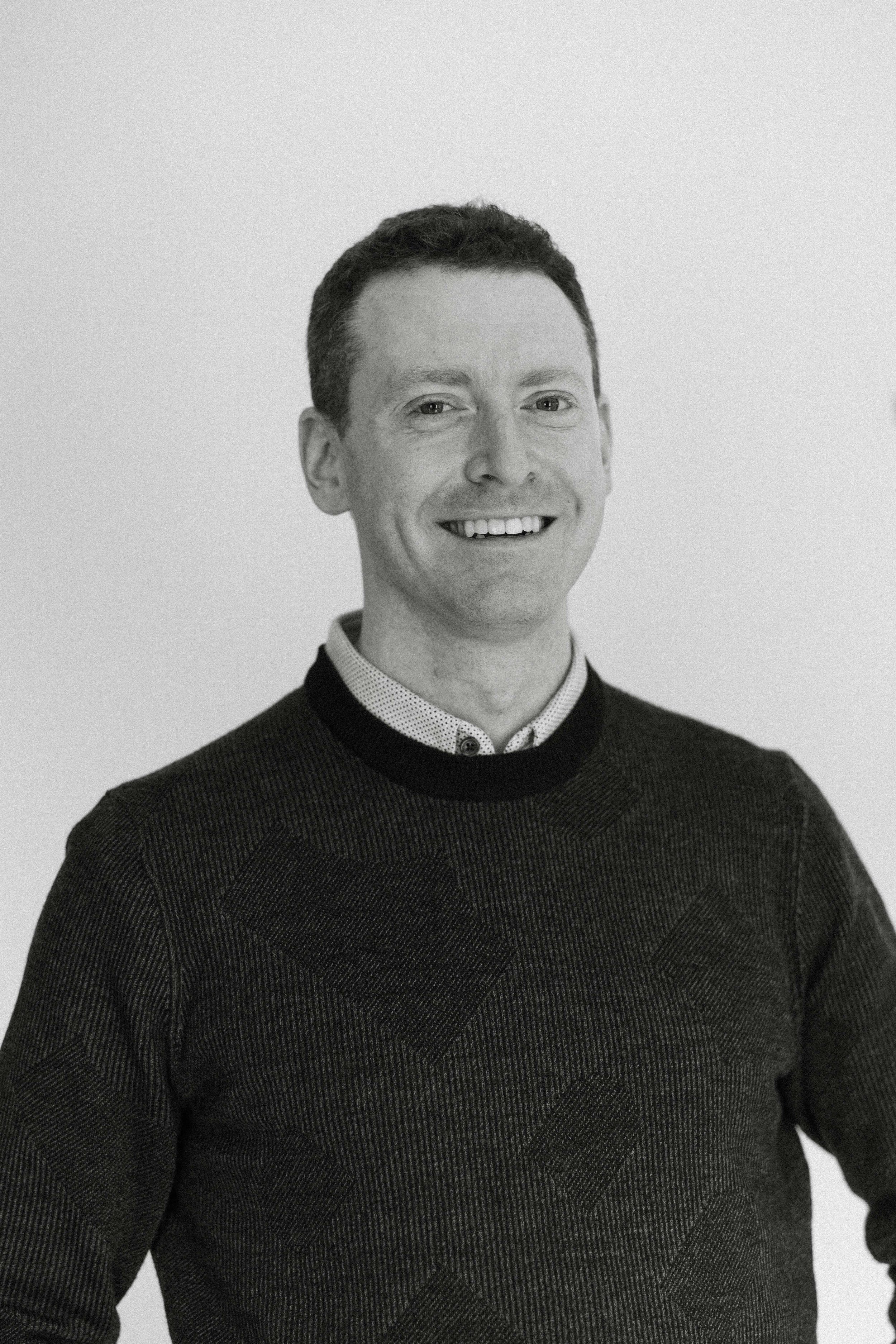 Black and white portrait of a smiling man with short hair, wearing a patterned sweater over a collared shirt, against a plain background.